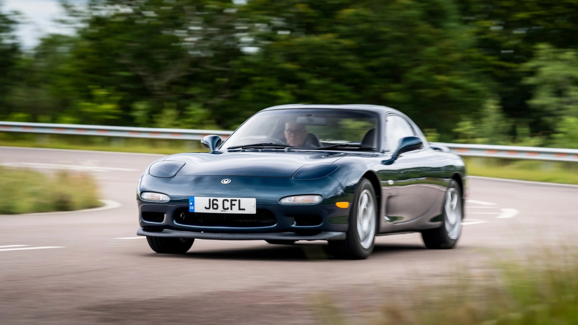 Dark green Mazda RX-7 sports car with pop-up headlights driving on a curved road.