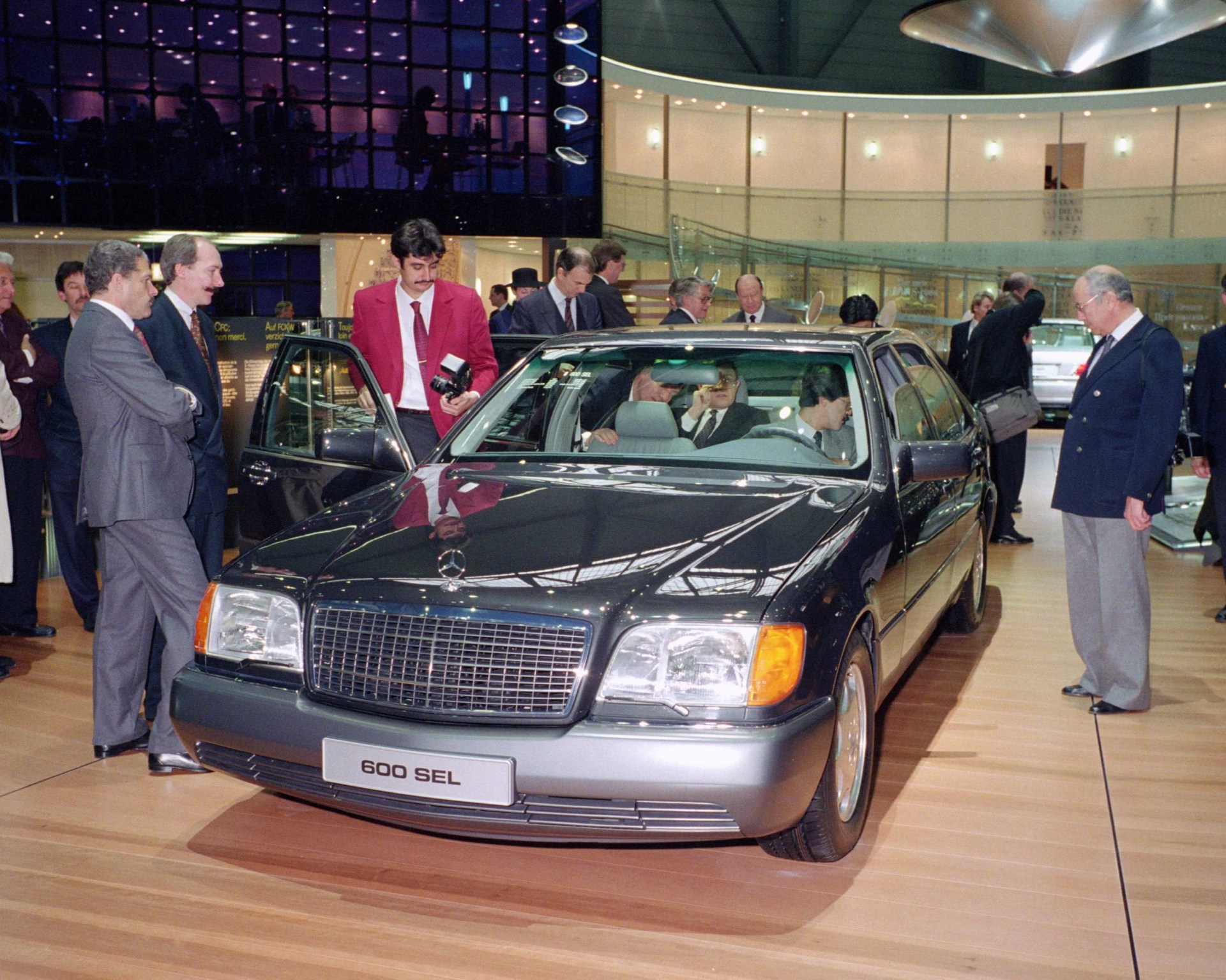 Black Mercedes-Benz 600 SEL sedan on display indoors with several people around it.