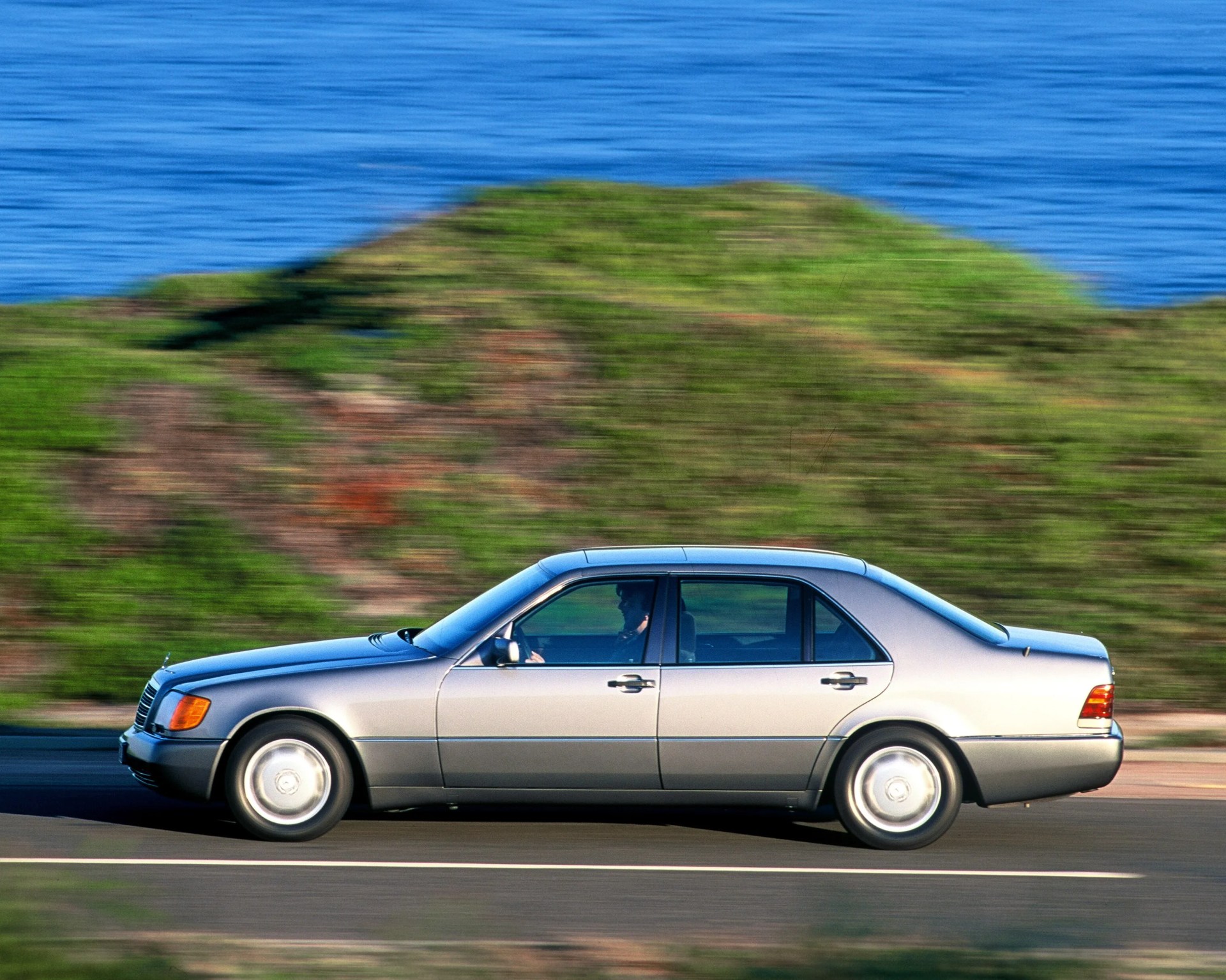 Silver four-door sedan driving on a road with blurred green hillside and blue water in the background.