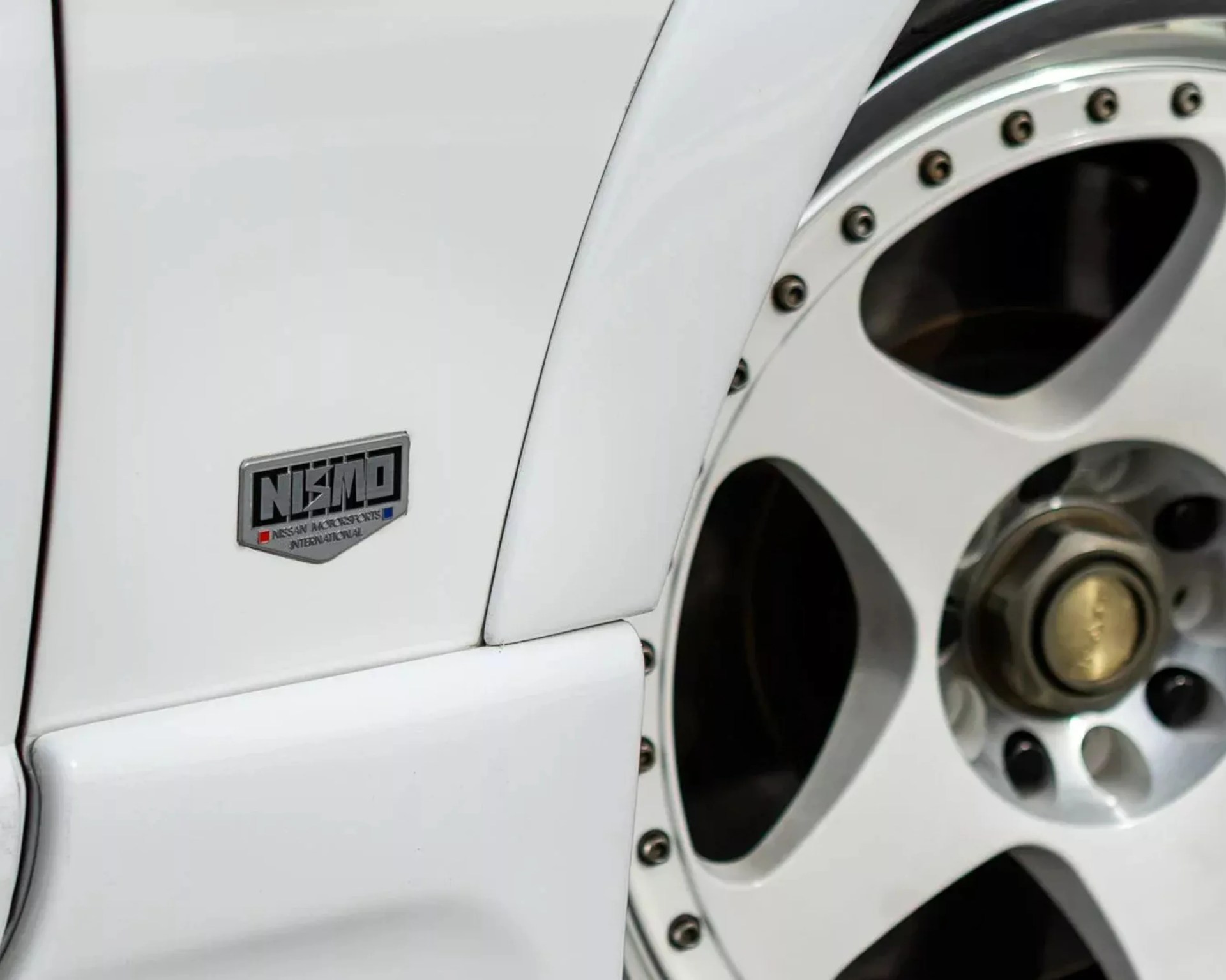 Close-up of a white car's fender with a NISMO badge and a silver multi-spoke alloy wheel.