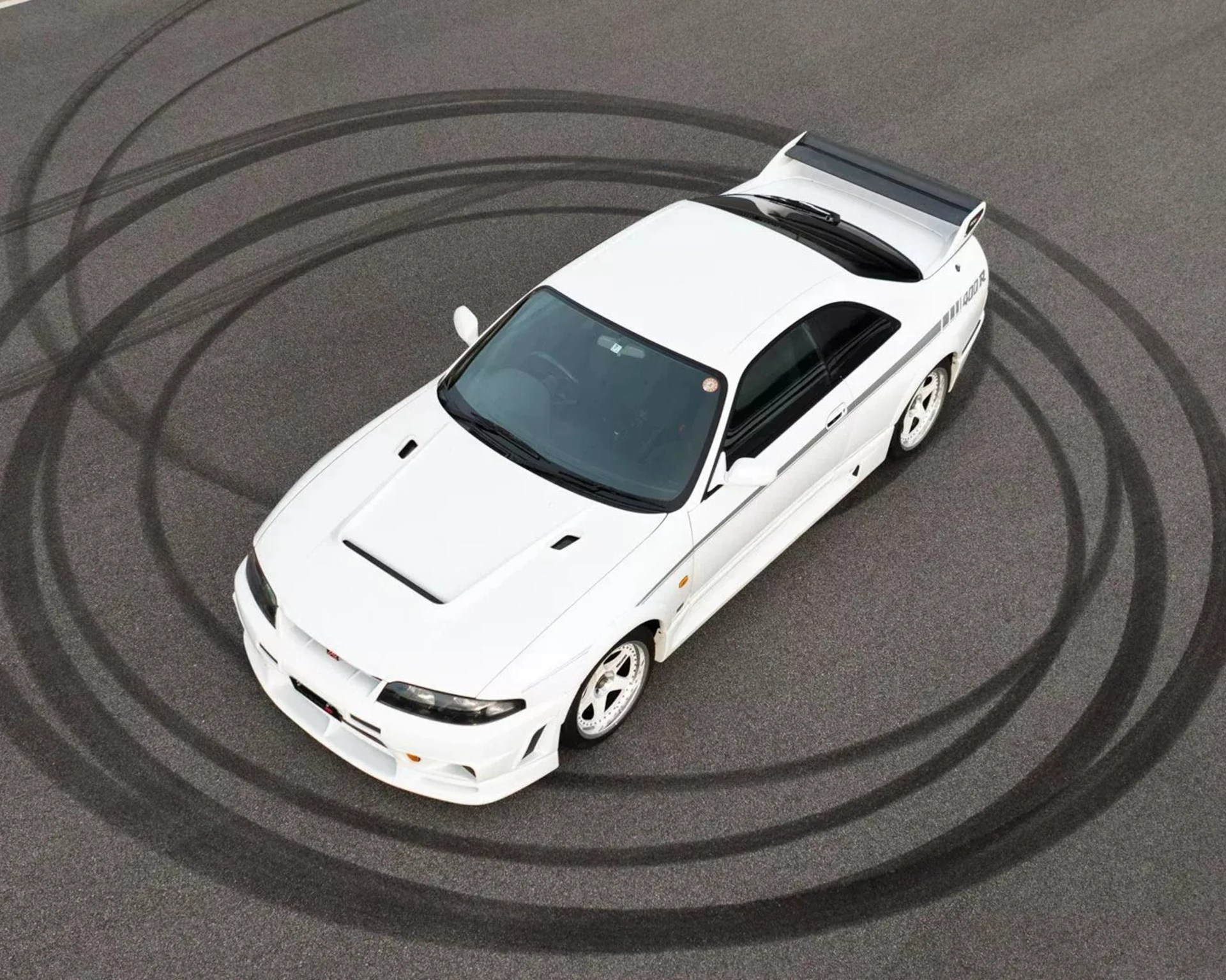 White Nissan Skyline GT-R R33 with black side stripe and rear spoiler on asphalt with tire skid marks.