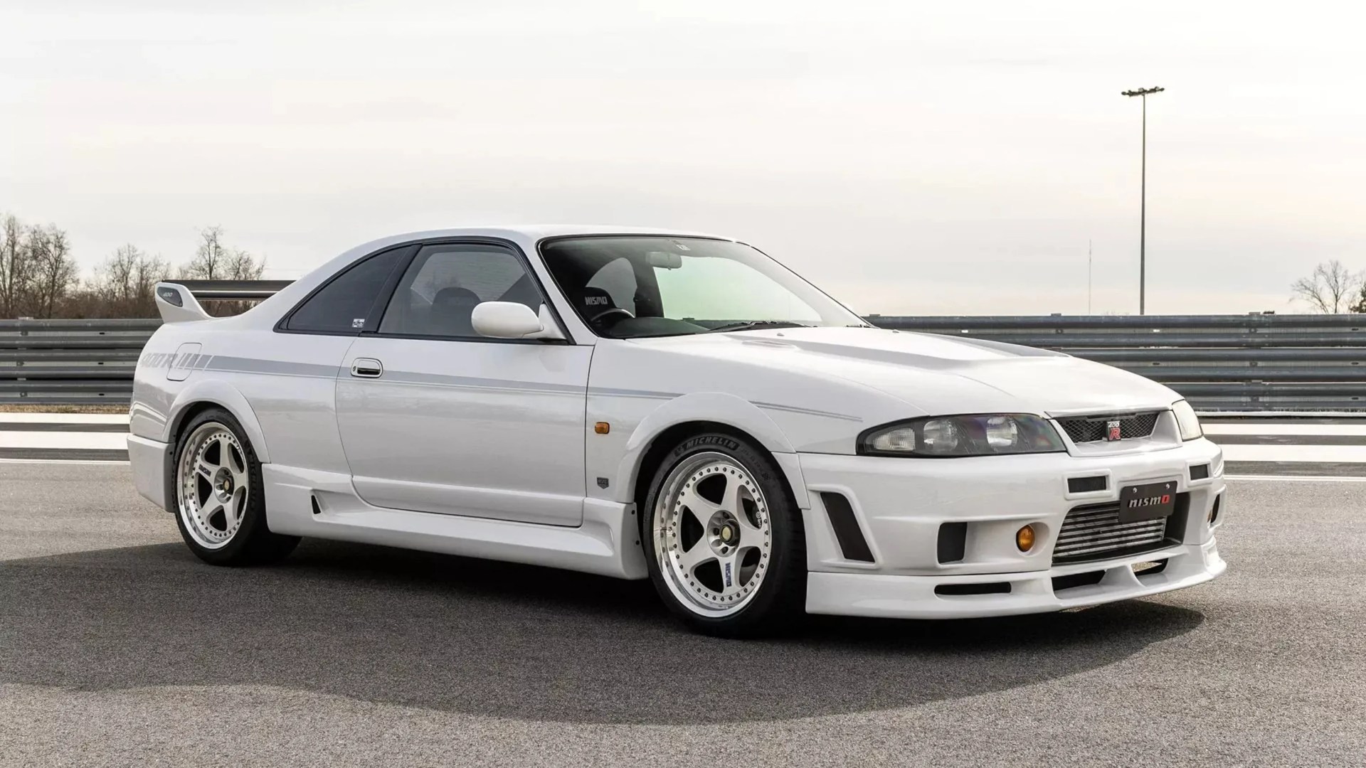 White Nissan Skyline GT-R R33 coupe with aftermarket wheels and rear spoiler on a racetrack.