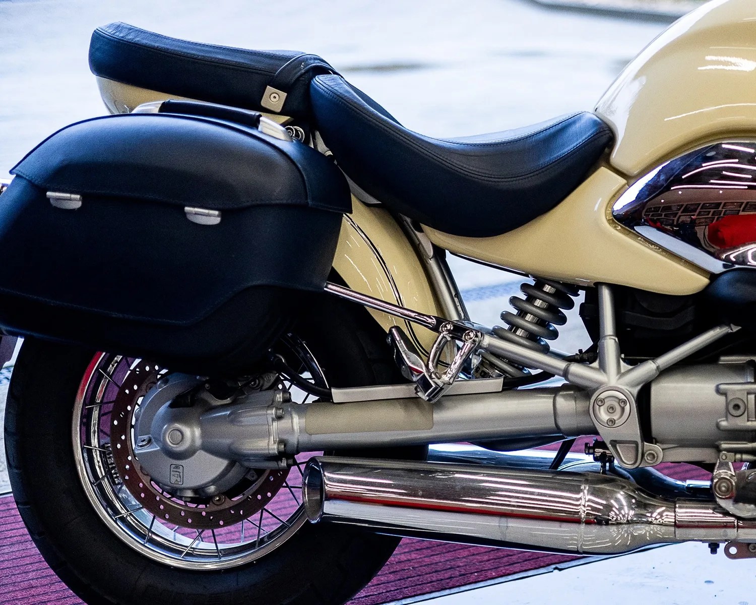 Close-up of a beige motorcycle's rear section featuring a black leather seat, black hard saddlebag, chrome exhaust, and rear suspension.