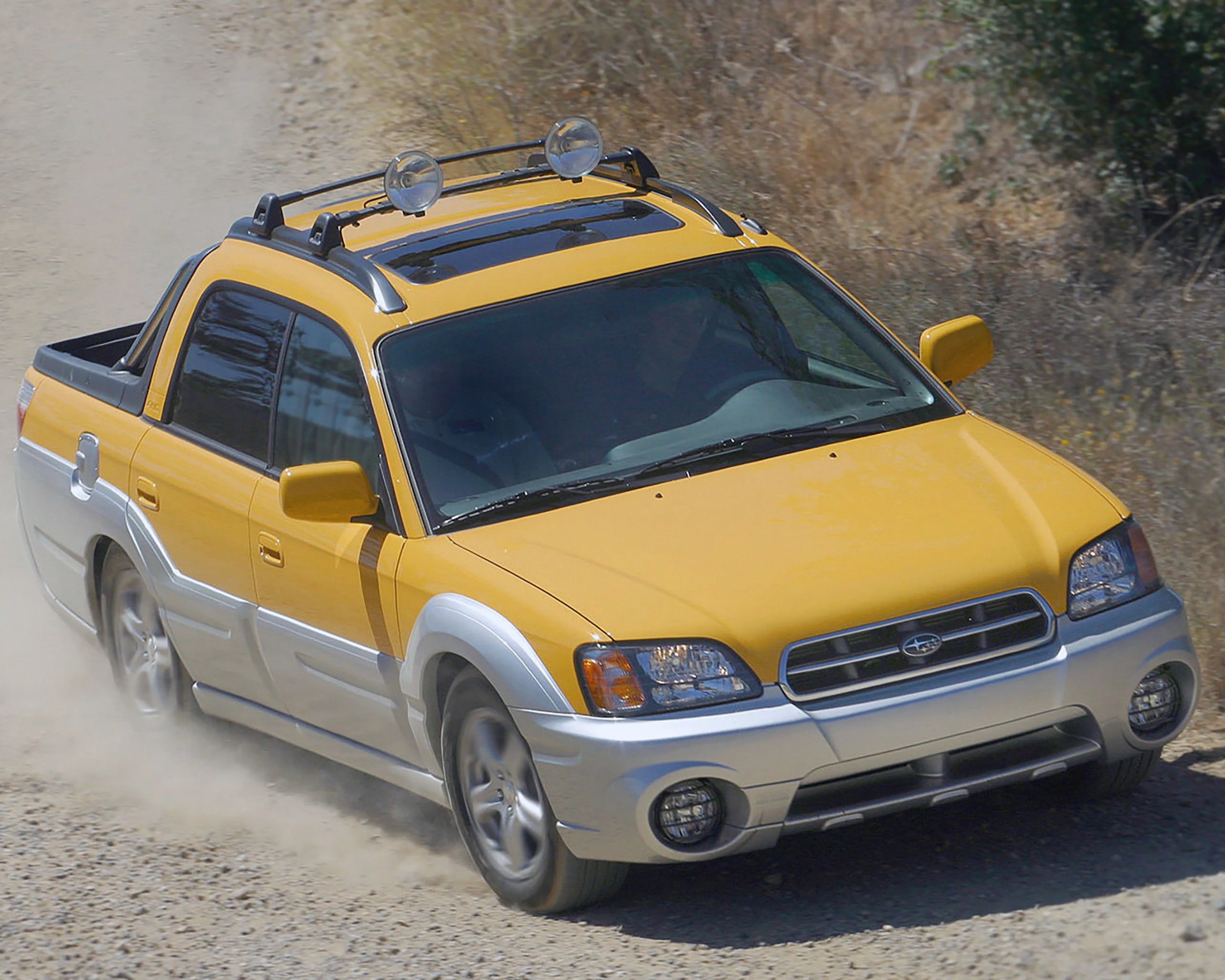 Yellow and silver Subaru Baja pickup truck driving on a dirt road with roof lights.