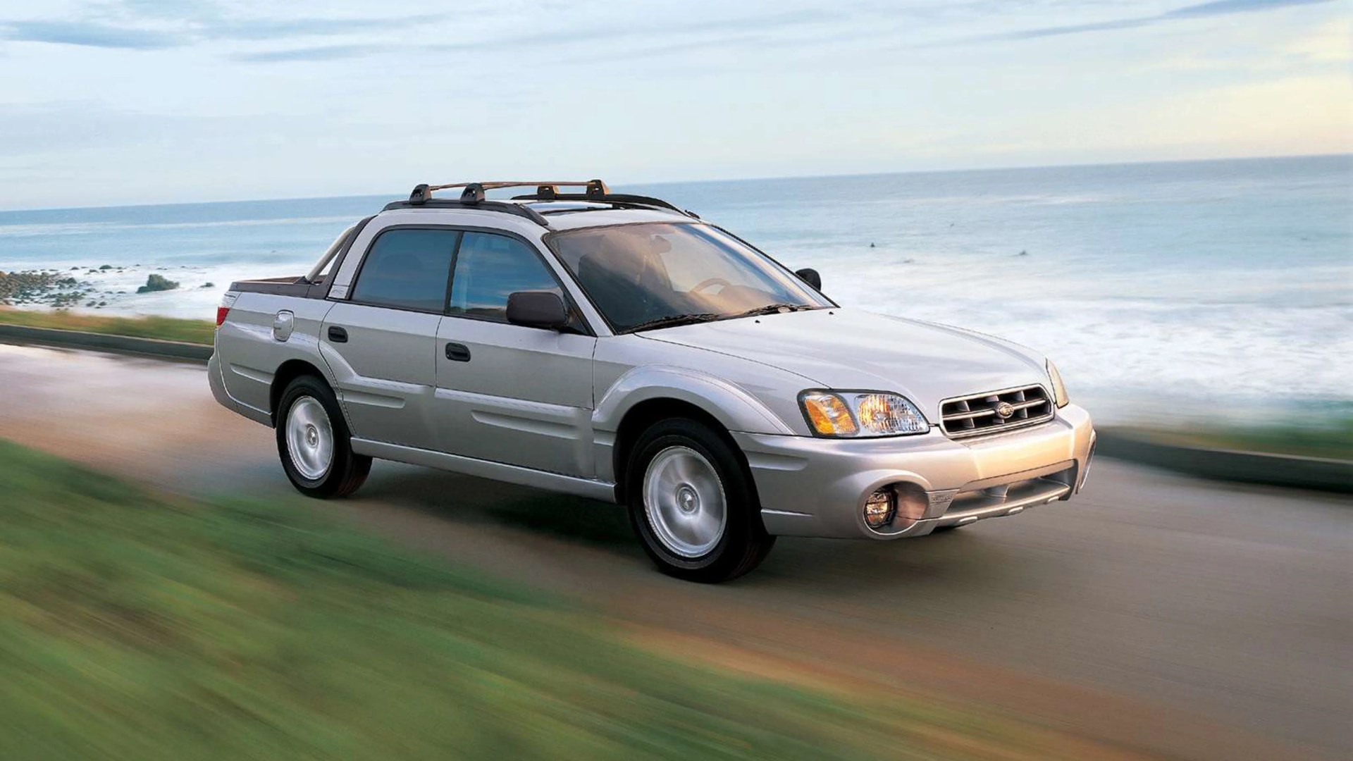 Silver Subaru Baja pickup truck driving on a coastal road with ocean in the background.