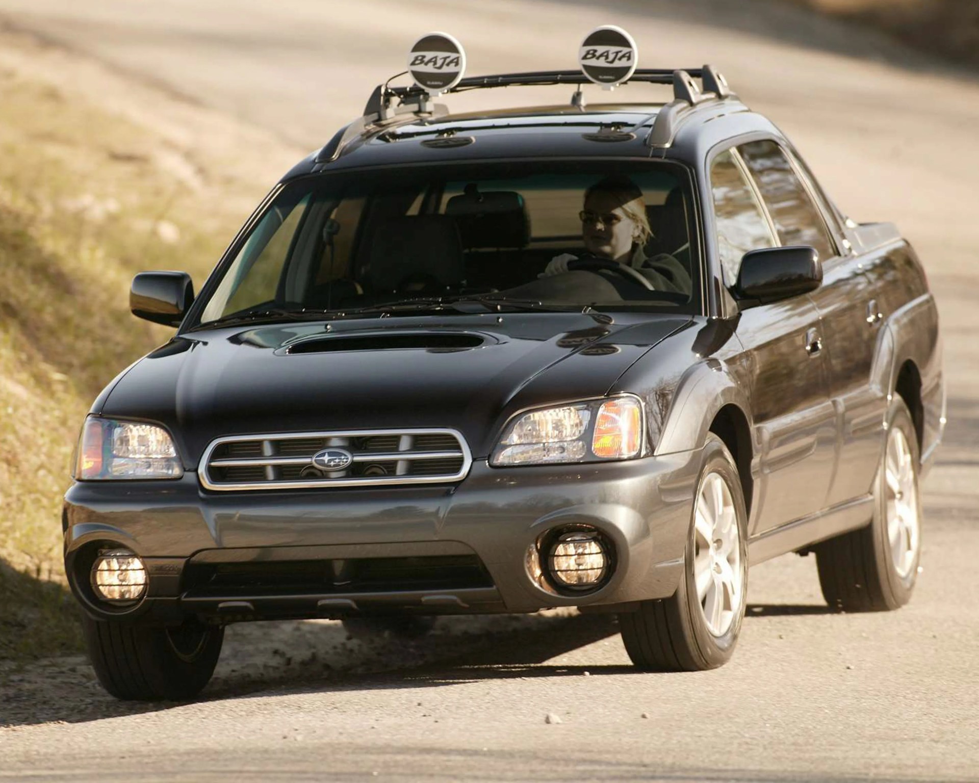 Black and gray Subaru Baja pickup truck with roof-mounted Baja lights driving on a dirt road.