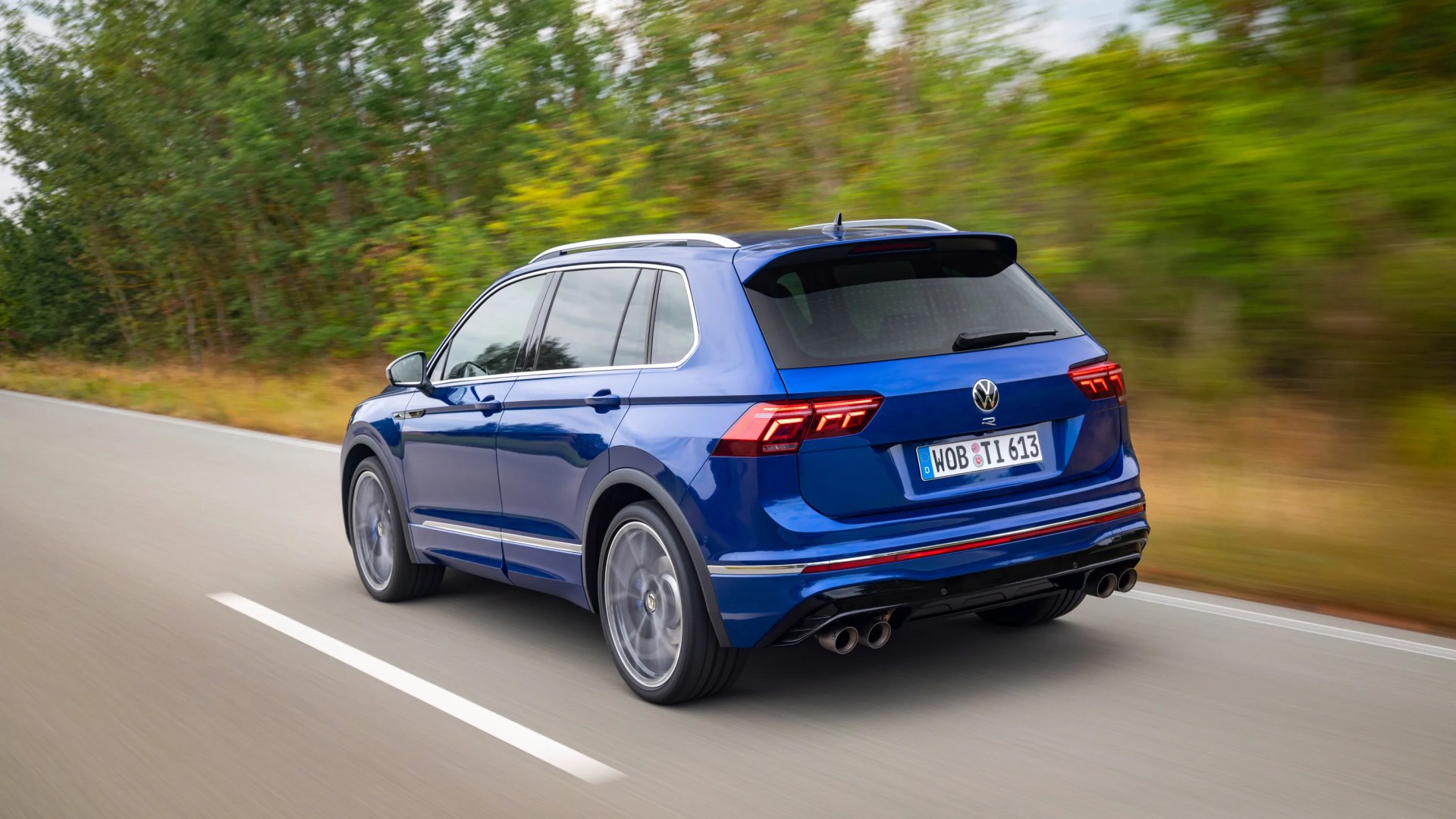 Blue Volkswagen SUV with tinted windows and quad exhaust pipes driving on a road with blurred trees in the background.