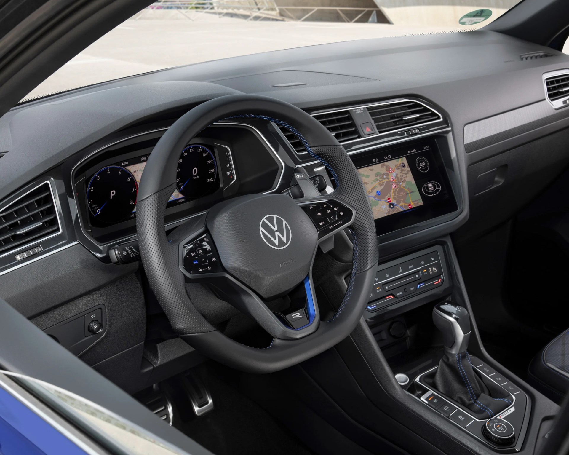 Black perforated leather Volkswagen steering wheel with blue stitching and digital dashboard behind it.