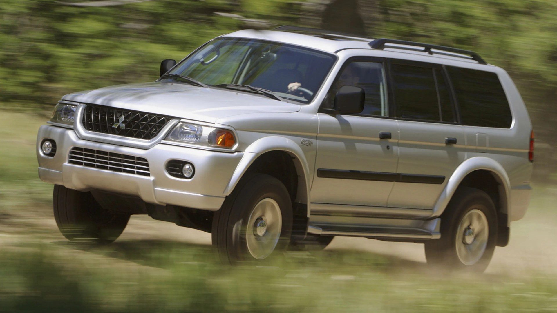 Silver Mitsubishi SUV with roof rails driving on a dirt road with blurred green background.