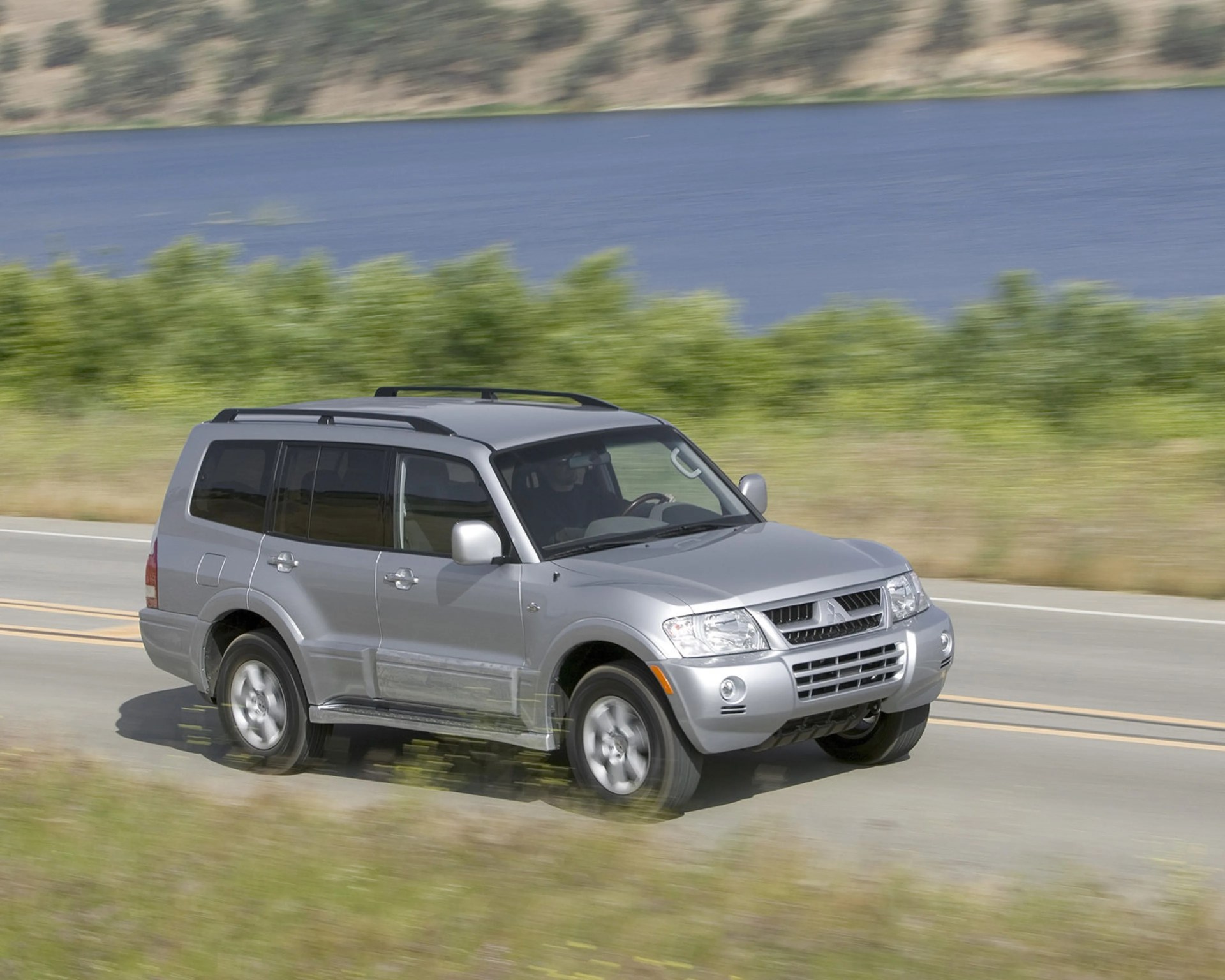Silver Mitsubishi Pajero SUV driving on a road with greenery and water in the background.