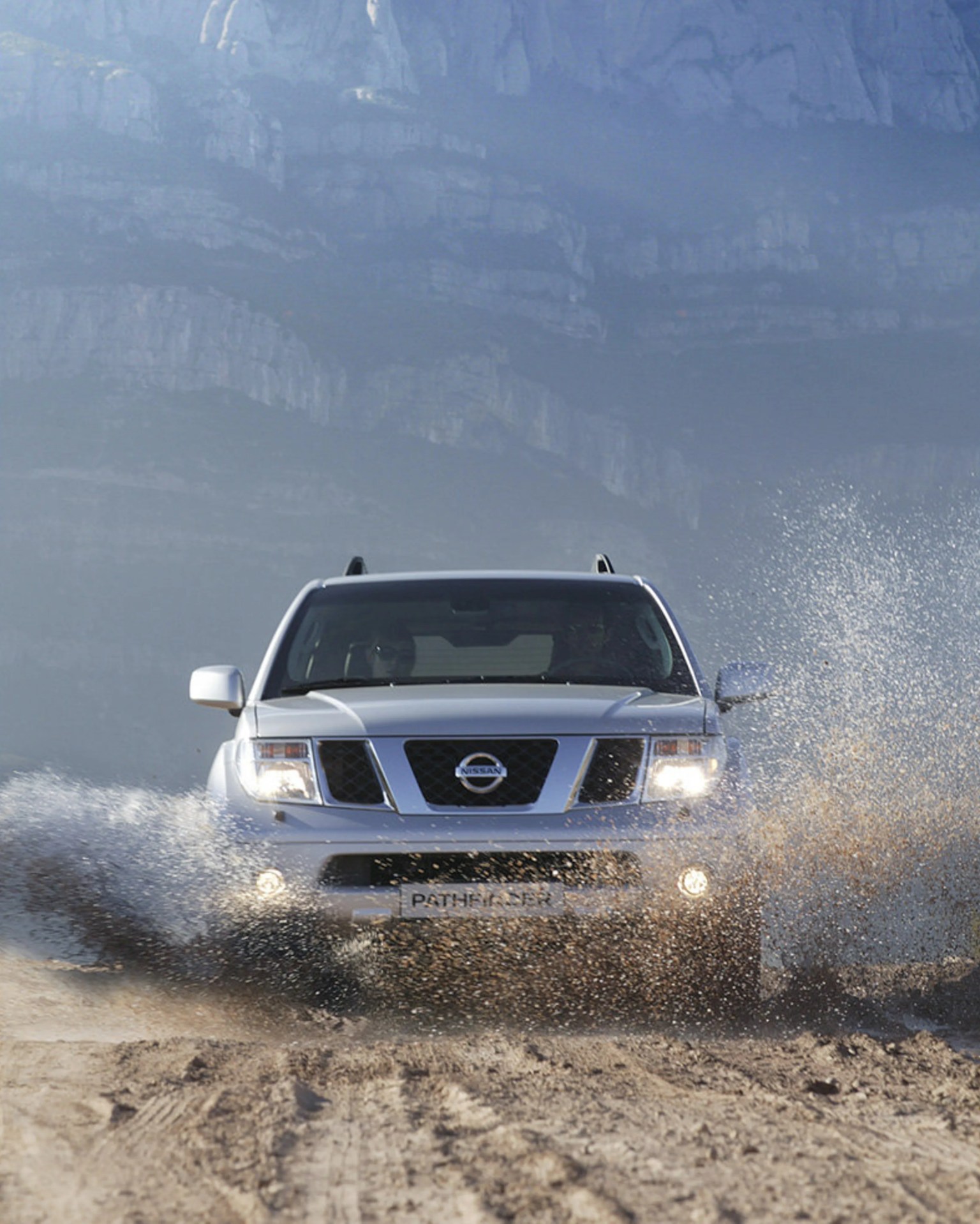 Silver Nissan Pathfinder SUV driving through muddy water with mountains in the background.
