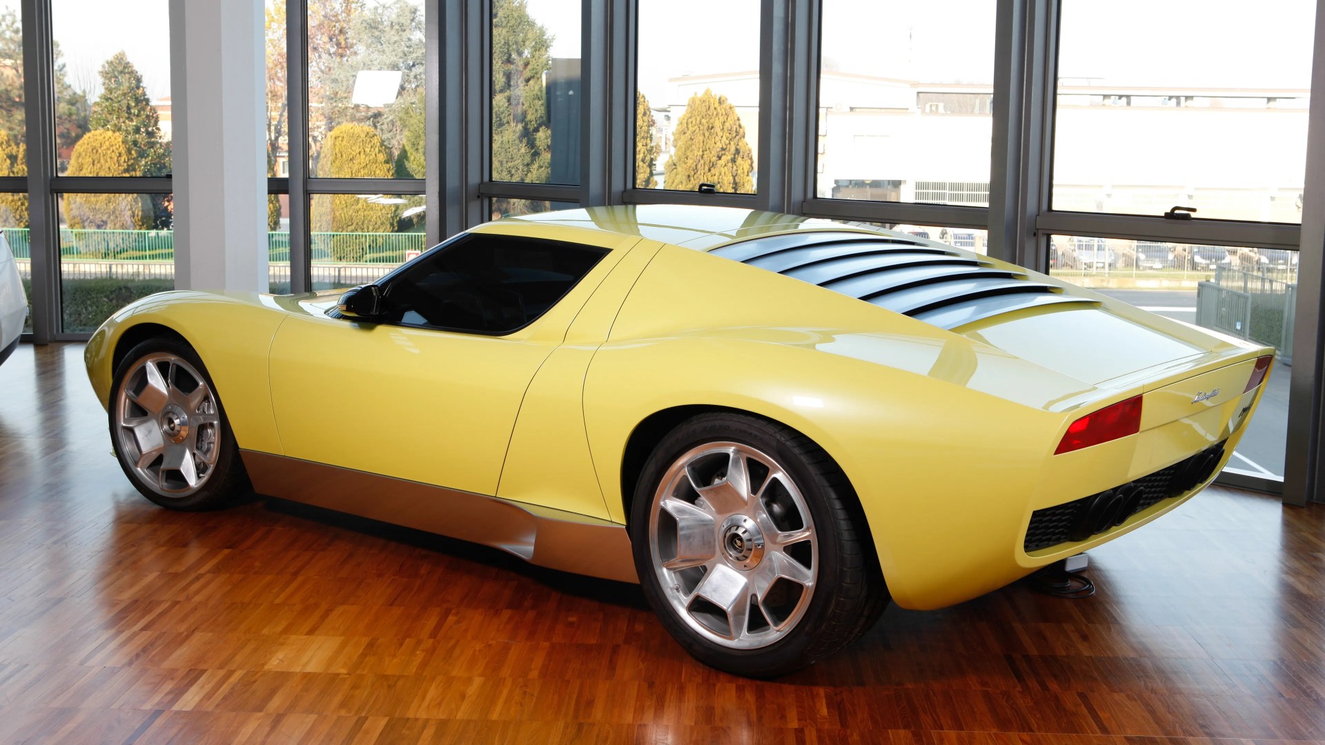 Yellow Lamborghini sports car with silver lower trim and rear window louvers parked indoors on wood floor.