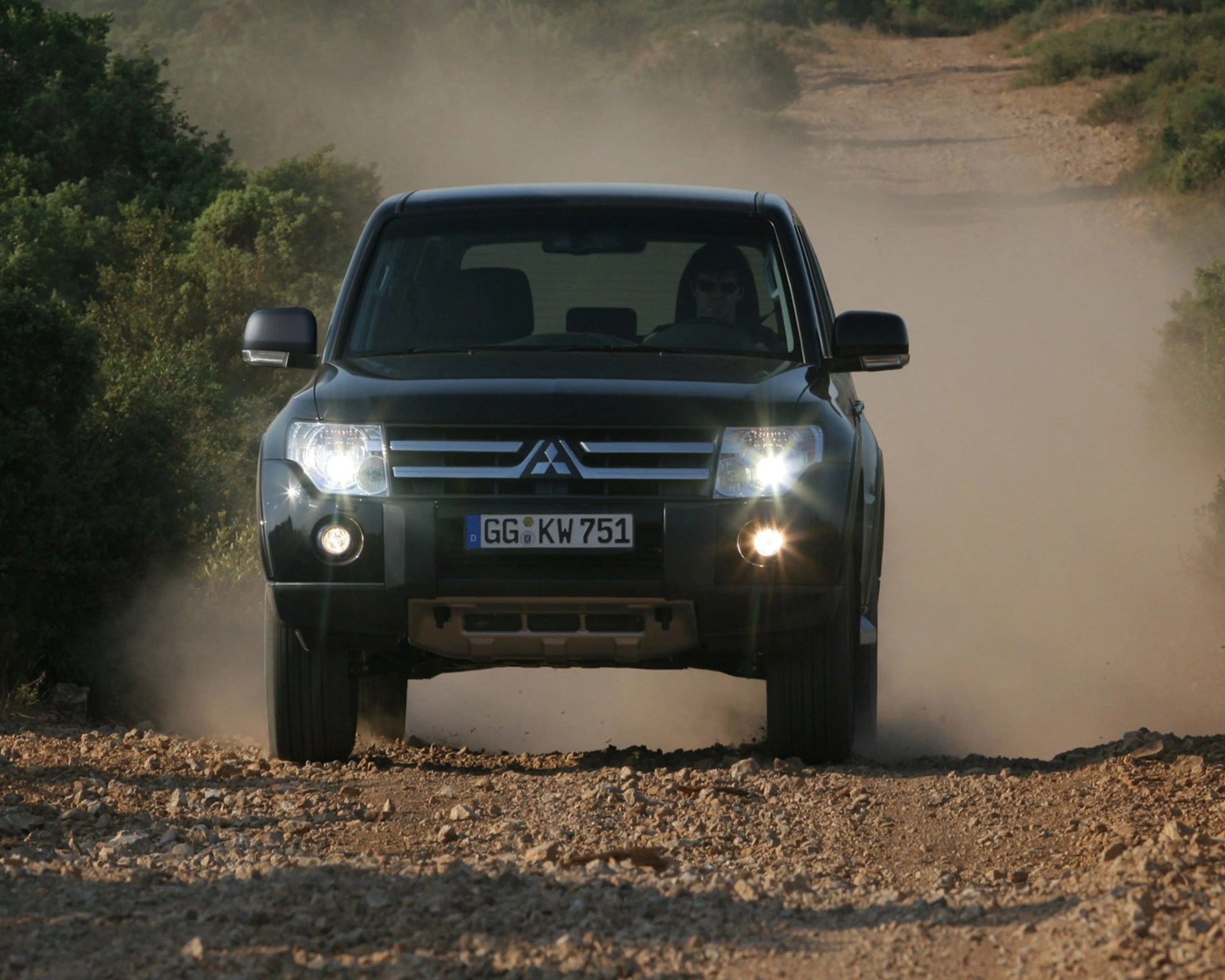 Black Mitsubishi SUV with headlights on driving on a rocky dirt road surrounded by greenery.