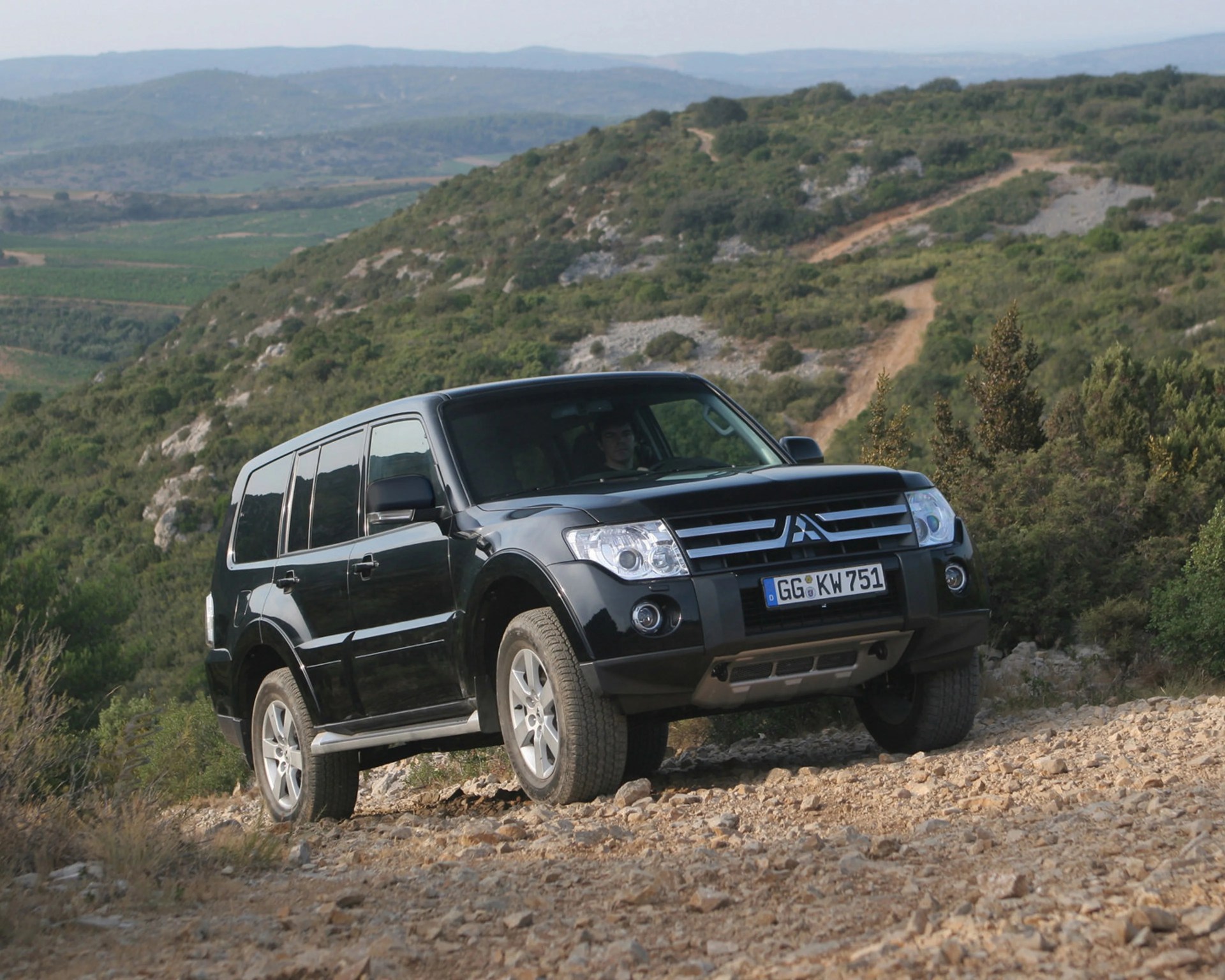 Black Mitsubishi SUV driving on a rocky dirt road in a hilly, green landscape.