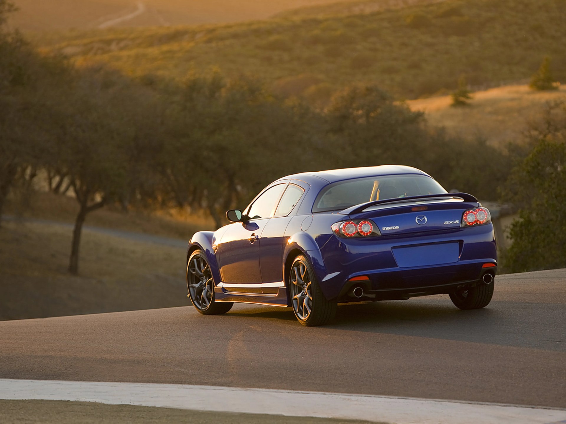 Blue Mazda RX-8 sports car parked on a road with trees and hills in the background at sunset.