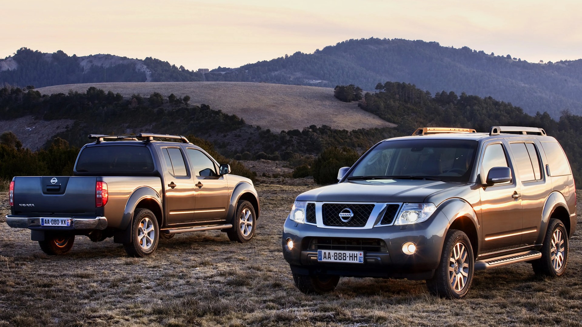 Two gray Nissan Navara pickup trucks parked on a grassy field with hills in the background at sunset.
