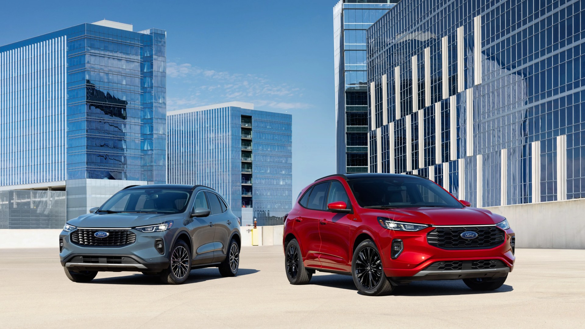 Two Ford SUVs, one gray and one red, parked on a rooftop with modern glass office buildings in the background.