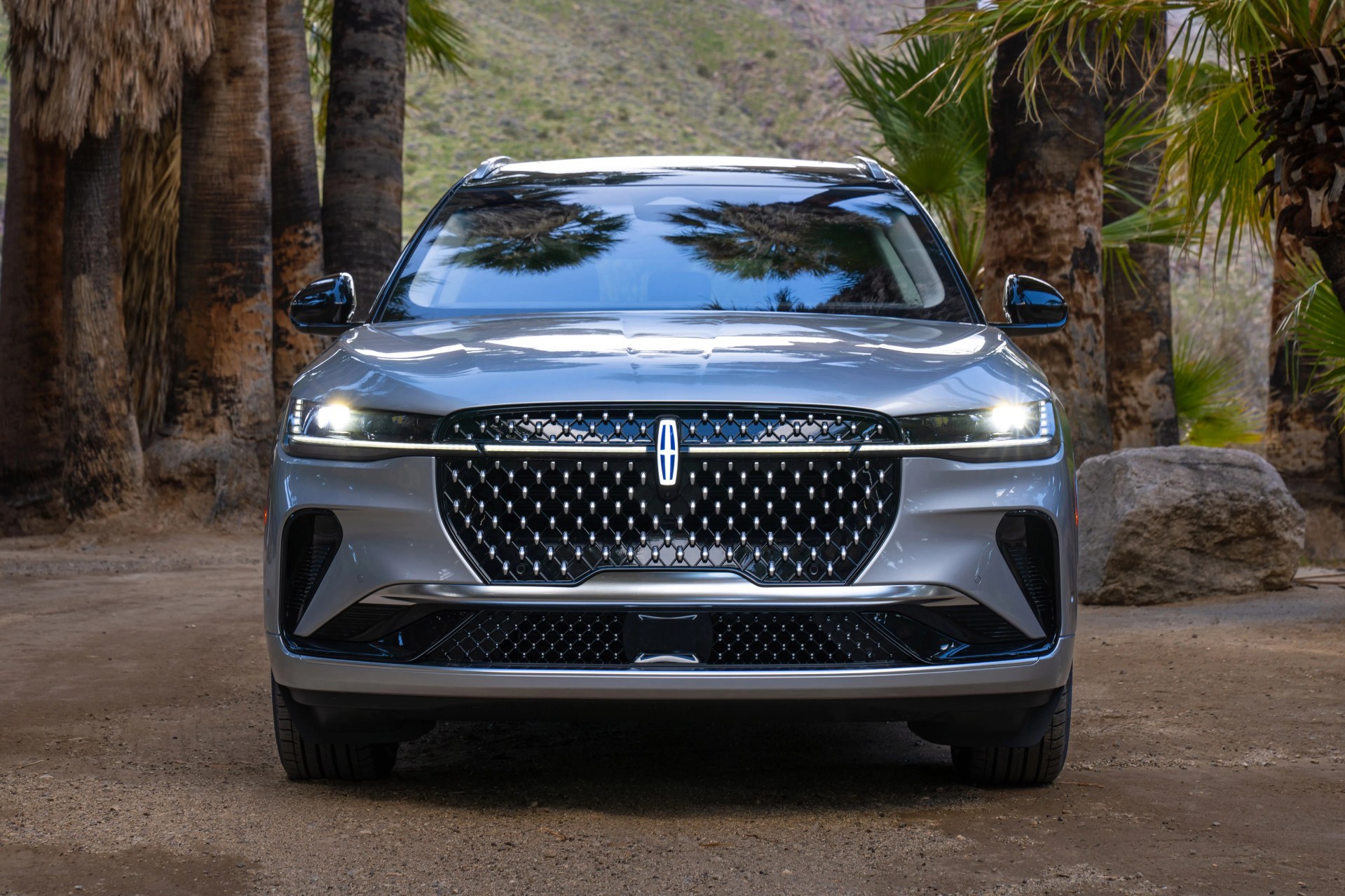 Front view of a silver Lincoln SUV with headlights on, parked on a dirt path surrounded by palm trees and rocks.