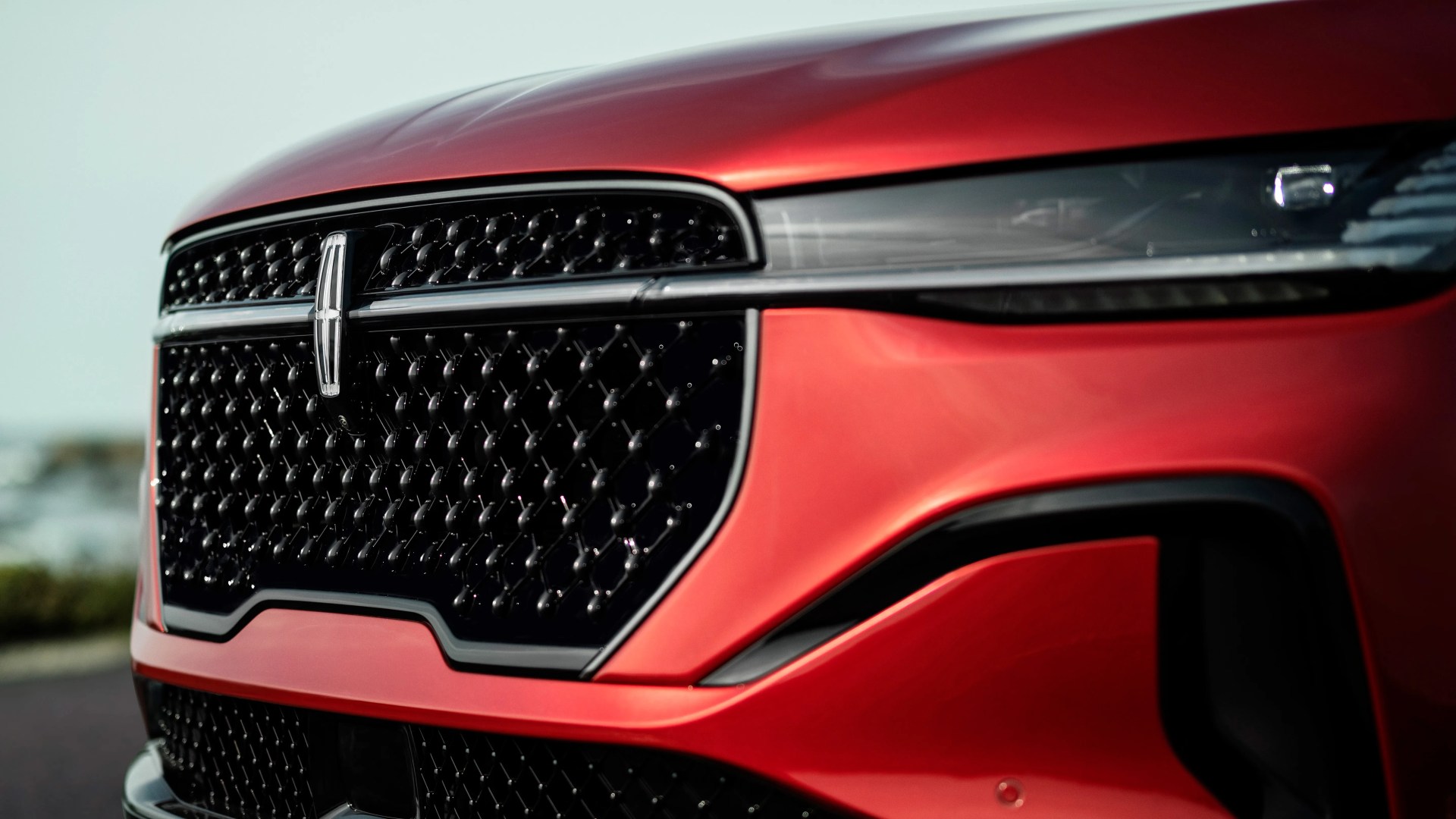 Front grille and headlight of a red Lincoln vehicle with a black mesh pattern and chrome emblem.