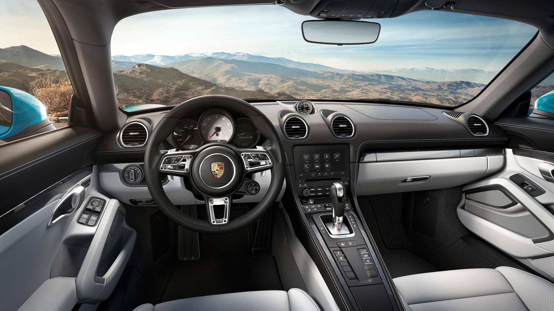 Interior view of a Porsche Cayman S with black and light gray leather, digital display, and a scenic mountain background.