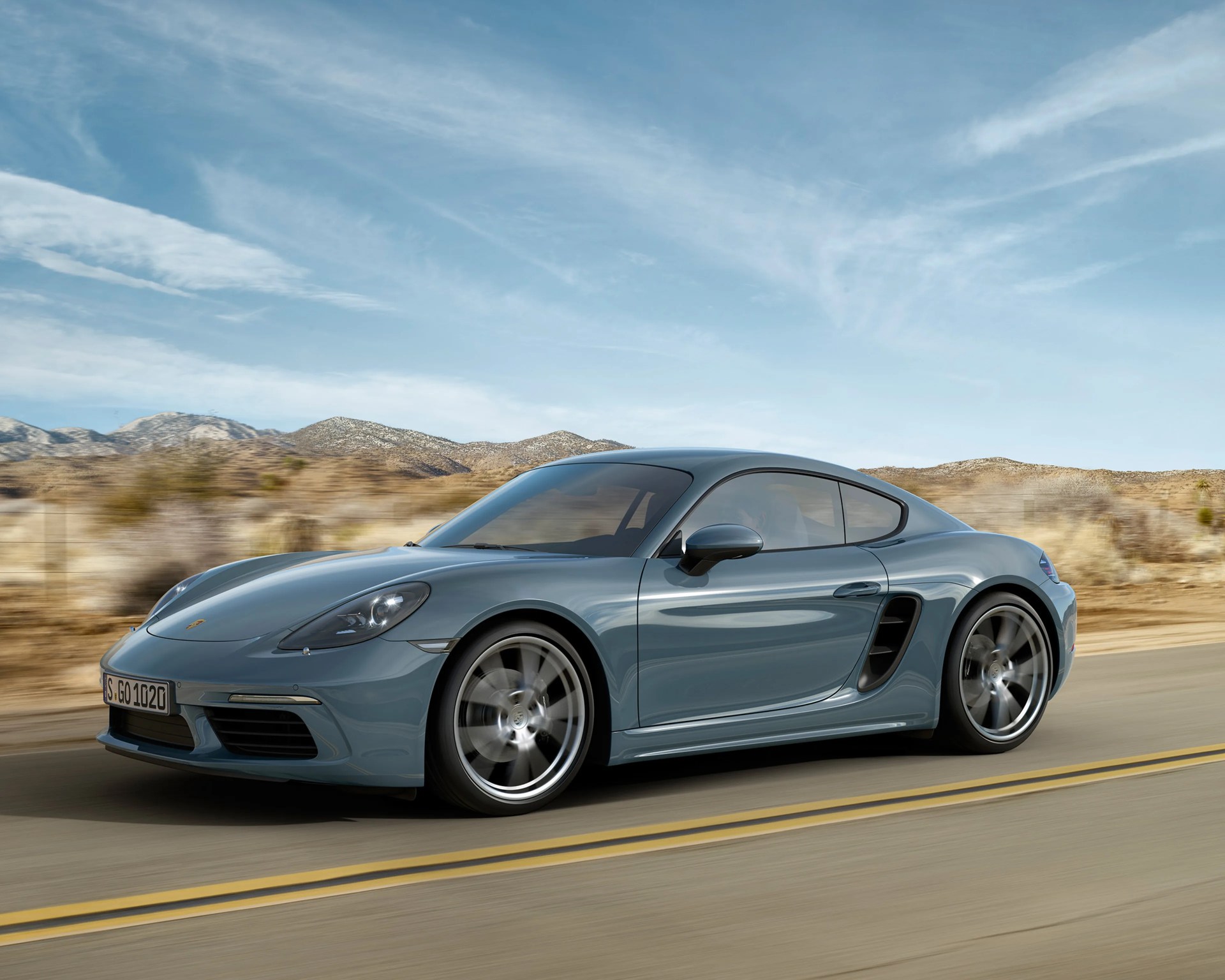 Blue-gray Porsche sports car driving on a desert road with mountains in the background.