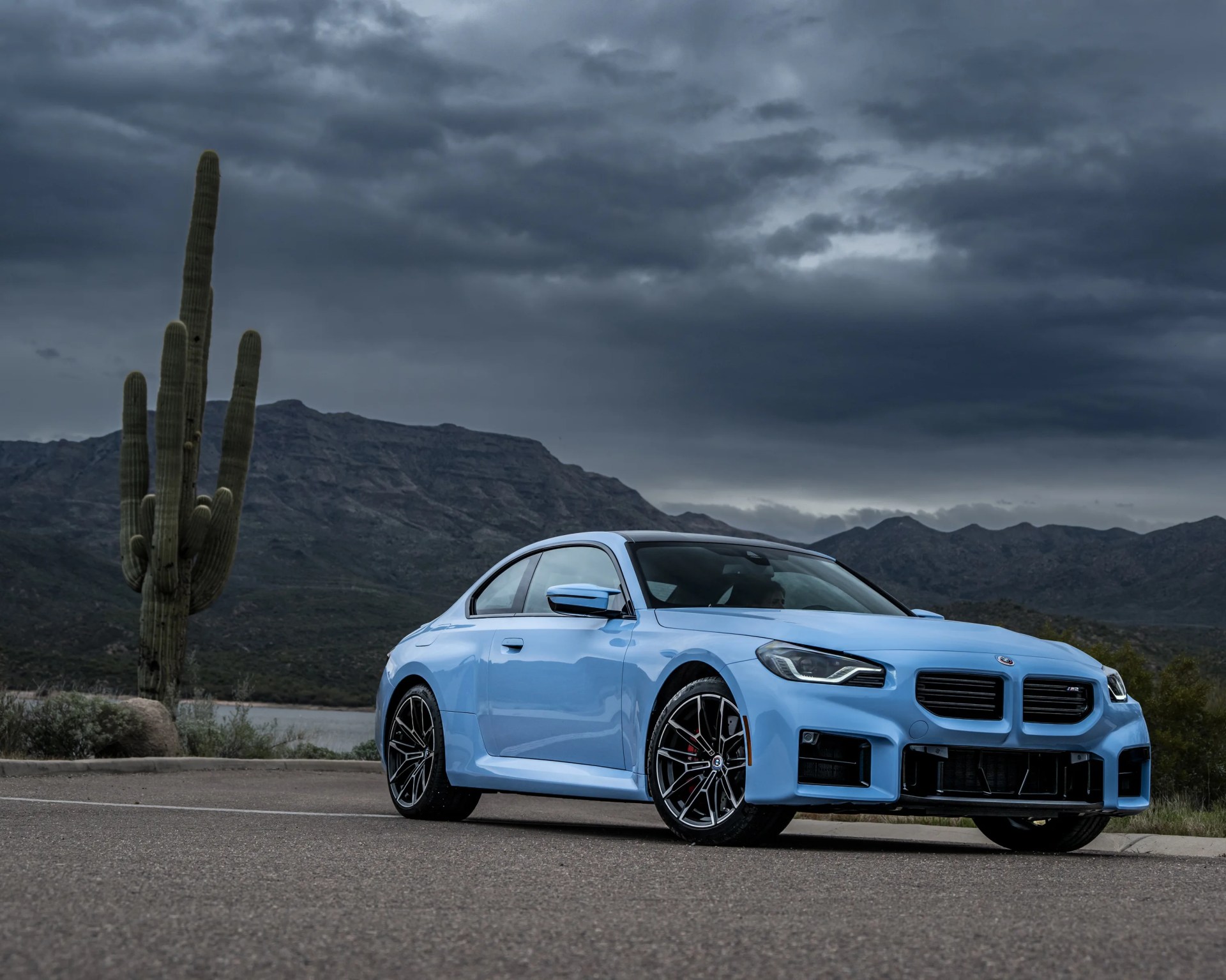 Light blue BMW M2 coupe parked on a road with mountains and a large cactus under a cloudy sky.