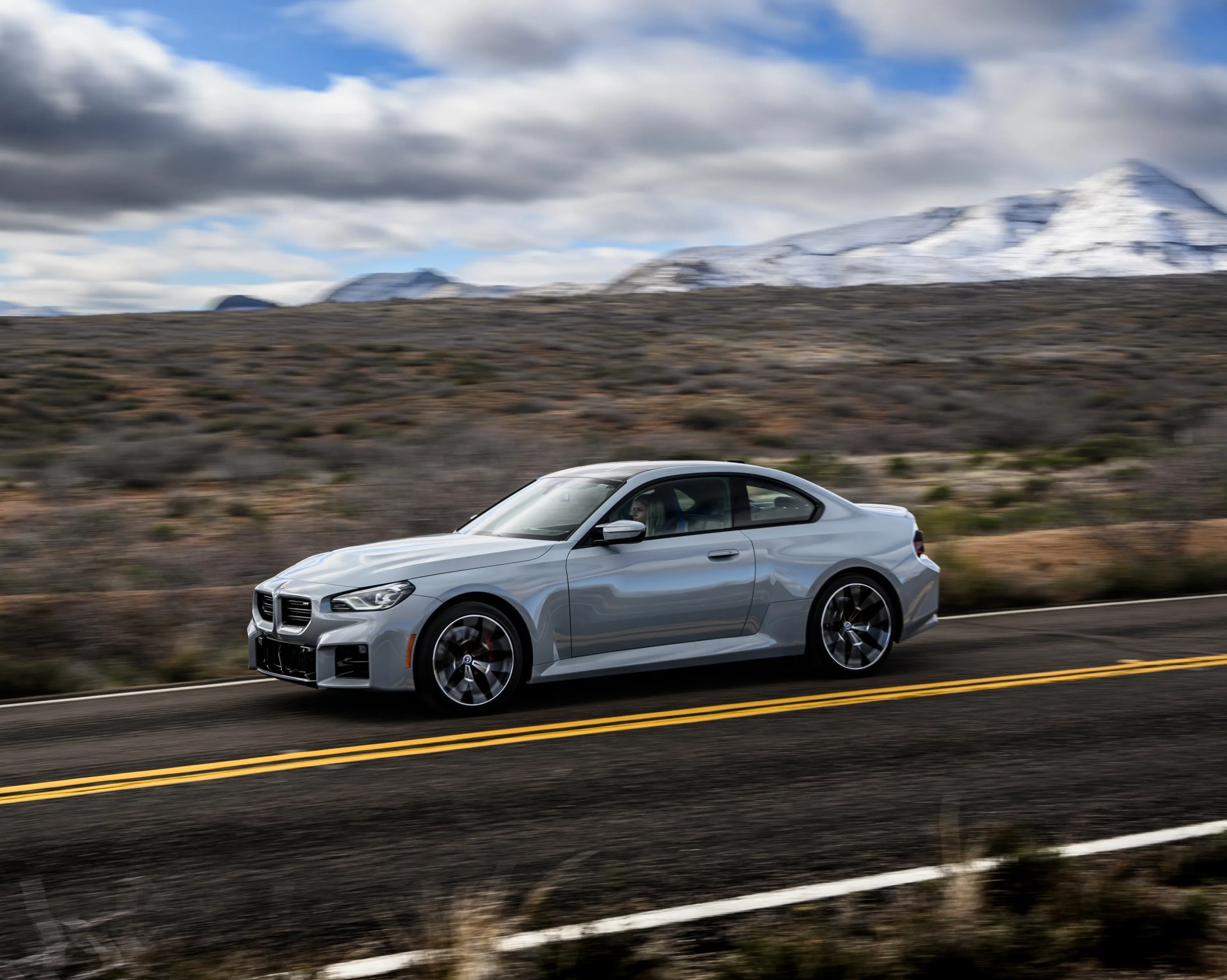 Silver BMW coupe driving on a two-lane road with mountainous landscape in the background.
