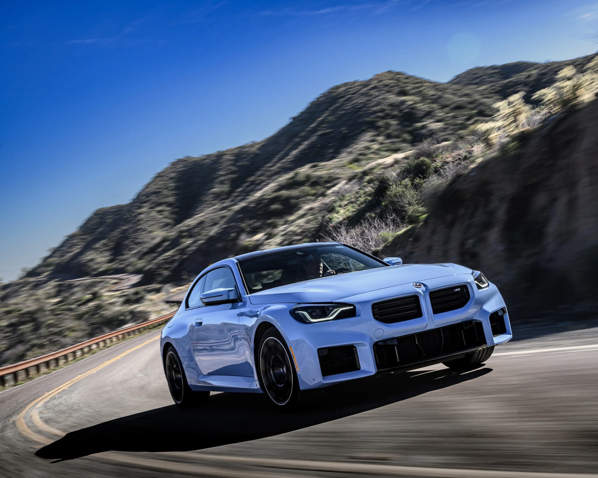 Light blue BMW M2 coupe driving on a winding mountain road under a clear blue sky.