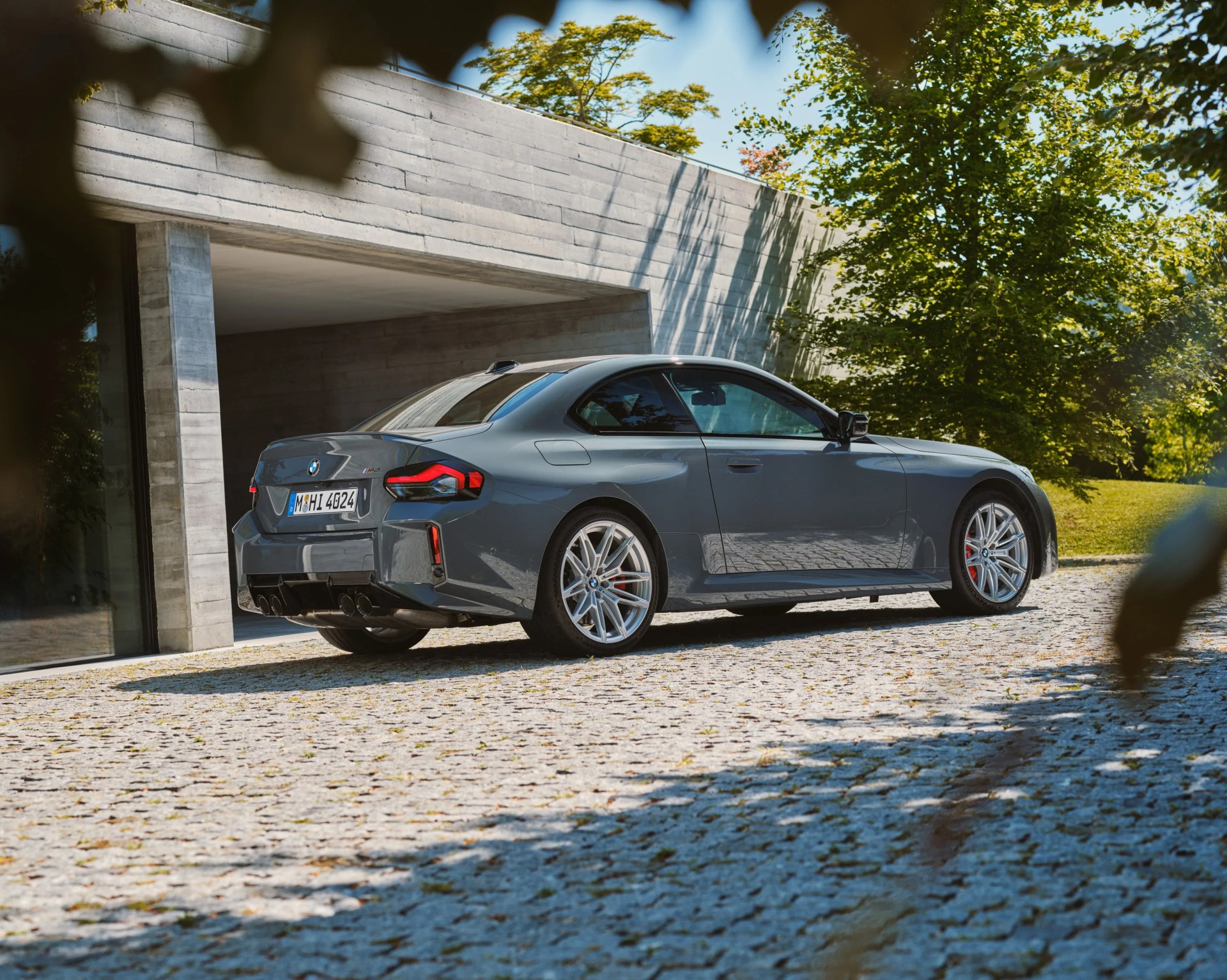 Gray BMW M2 coupe parked on a cobblestone driveway near a modern concrete building with green trees in the background.