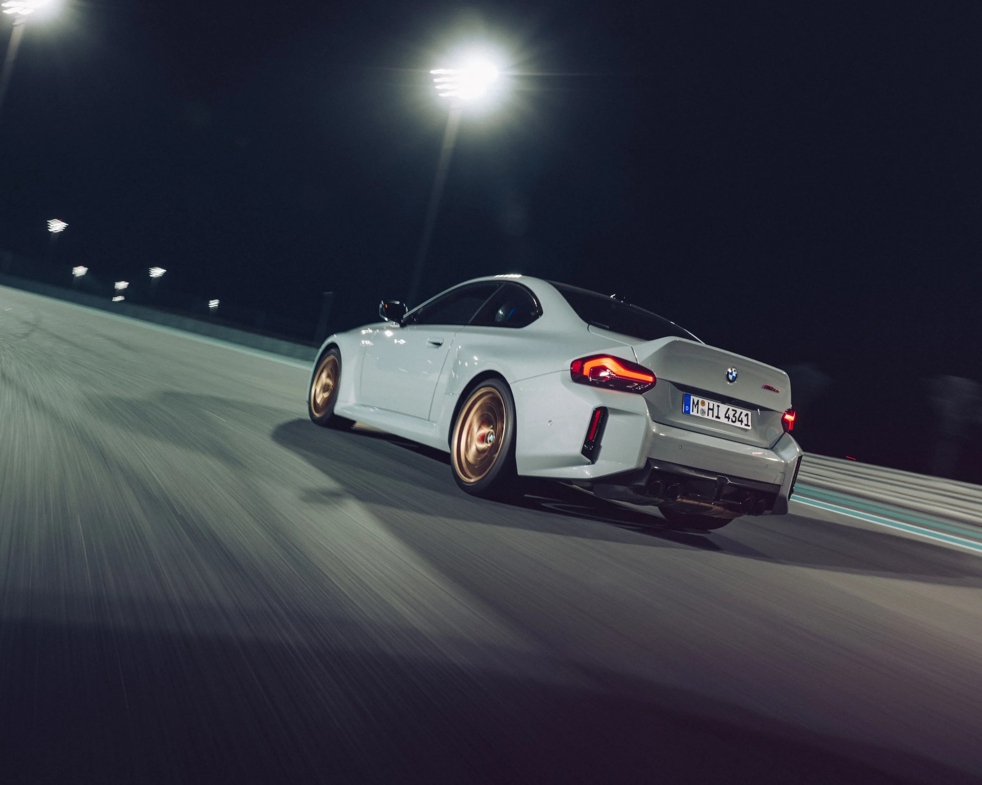 White BMW M4 coupe with gold wheels driving on a lit racetrack at night.