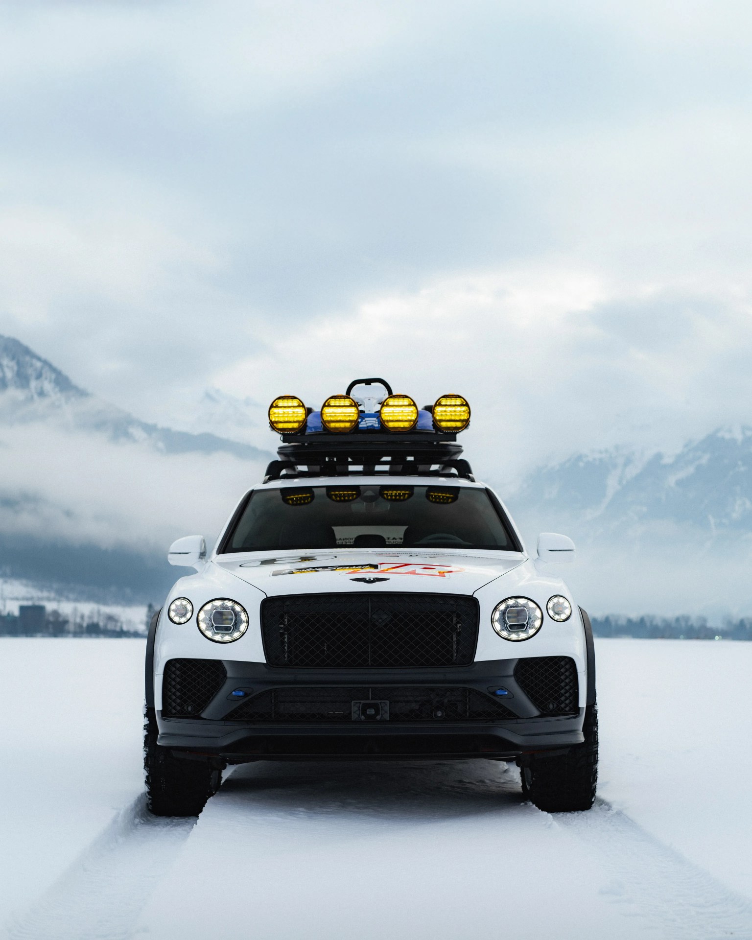 White Bentley SUV with roof-mounted yellow off-road lights on a snowy landscape with mountains in the background.