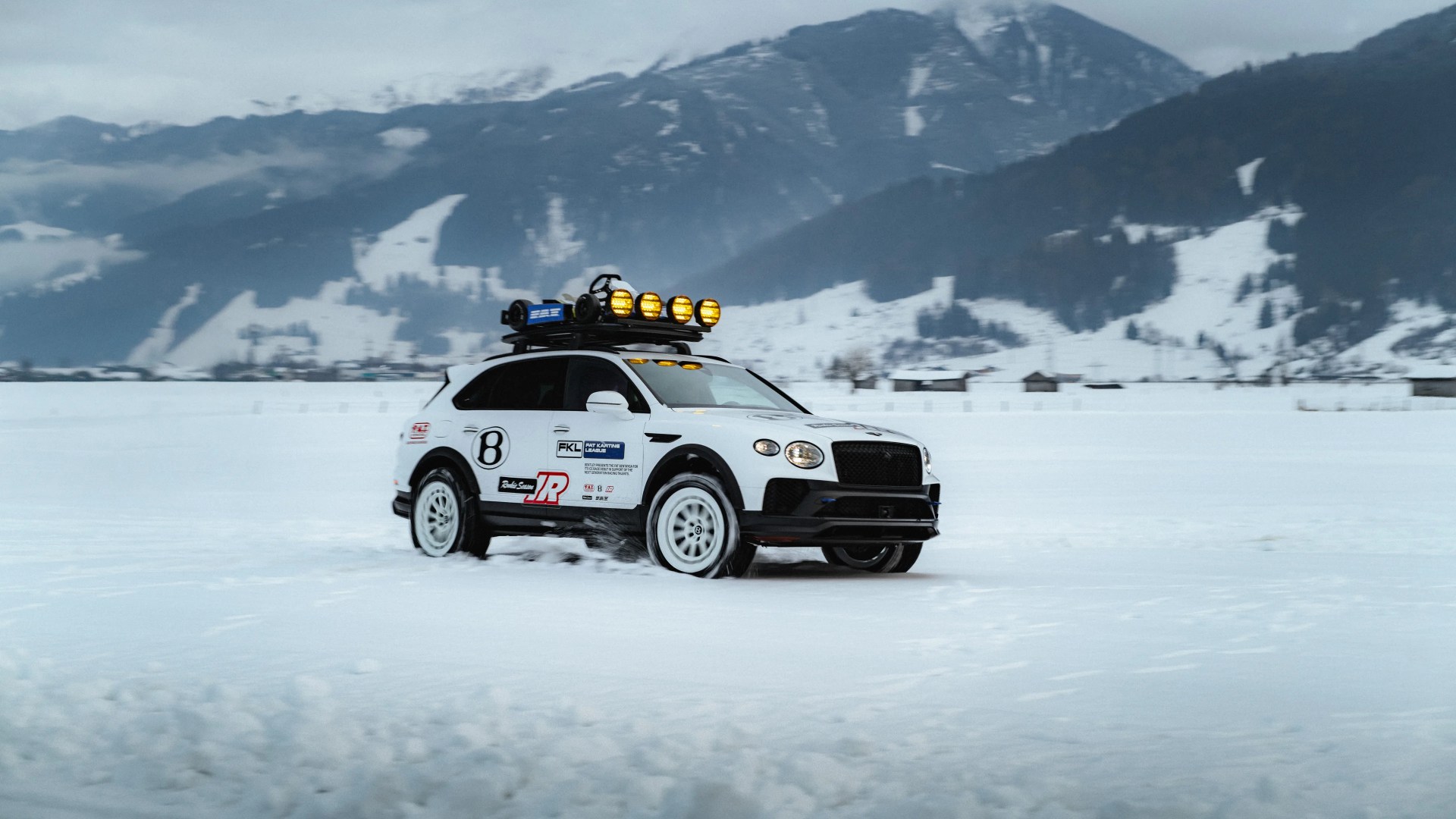 White rally SUV with roof lights and decals driving on snow with mountains in the background.