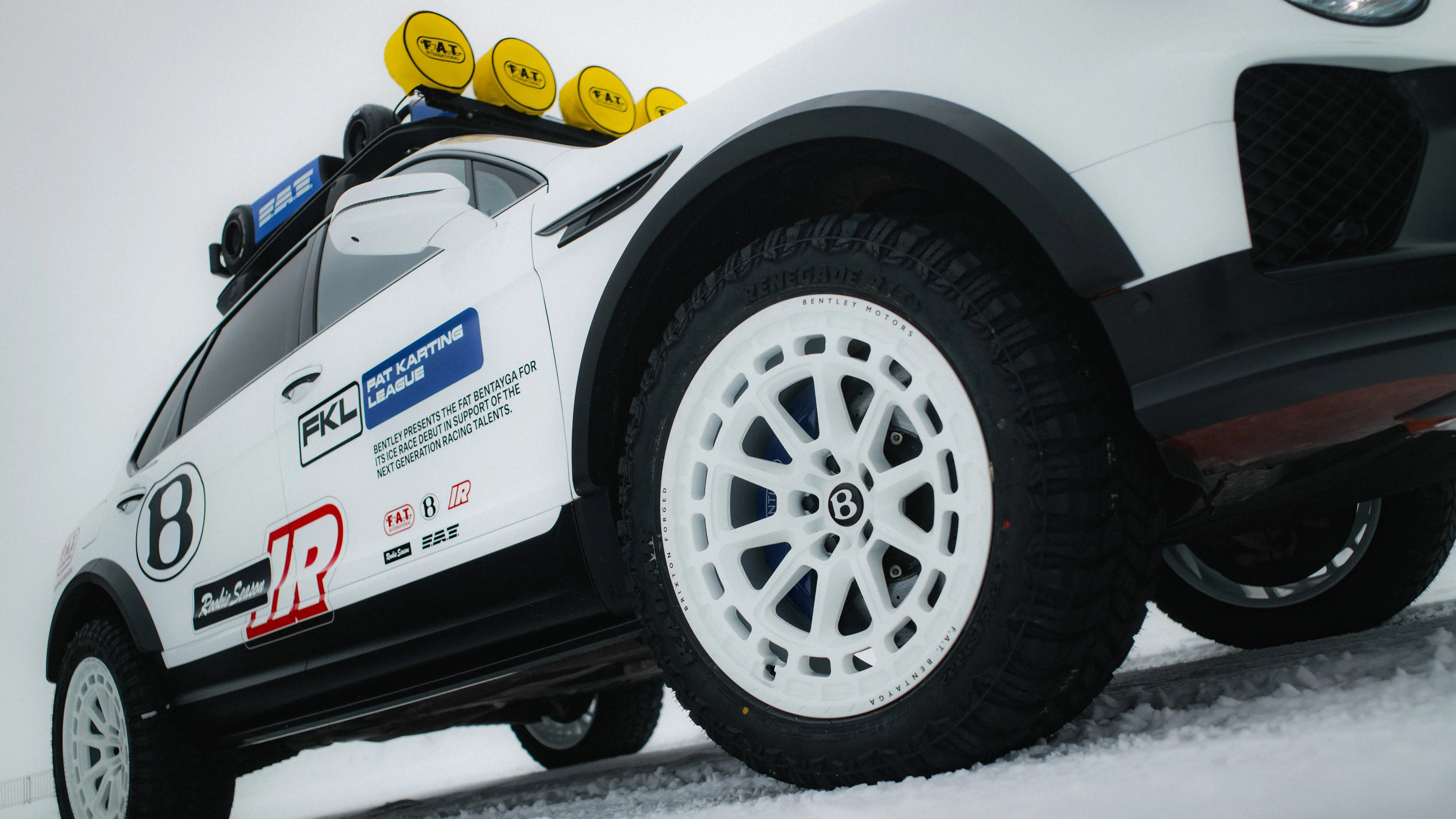 White Bentley Bentayga SUV with off-road tires, roof rack with yellow lights, and racing decals on snow.