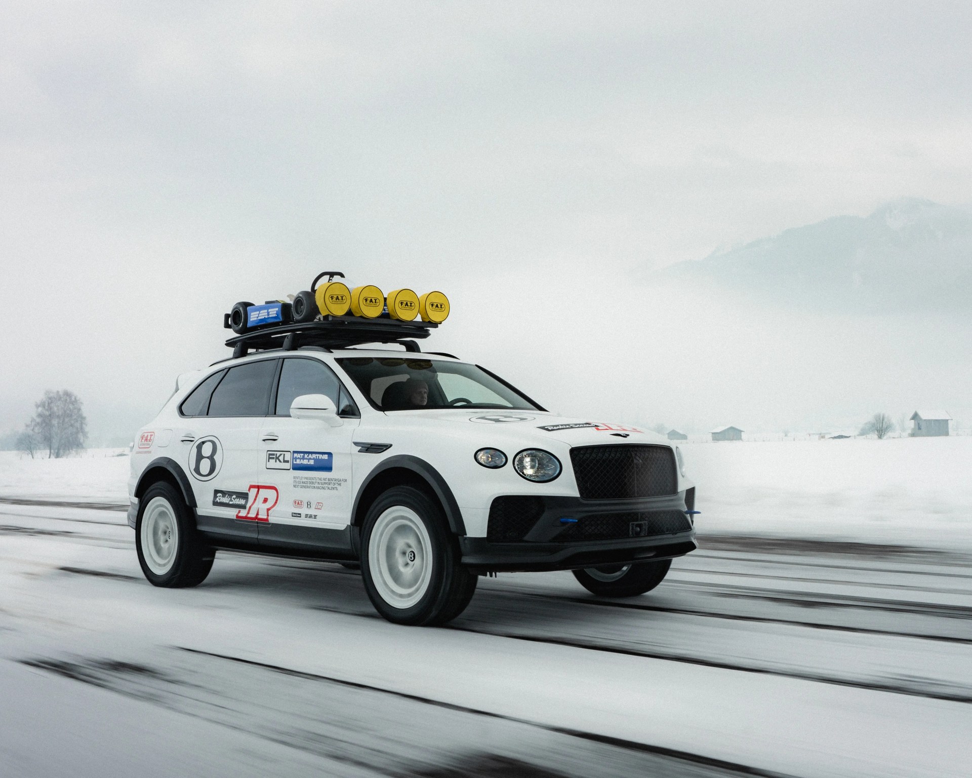 White rally-style SUV with black trim, roof rack with yellow lights, and racing decals driving on a snowy road.