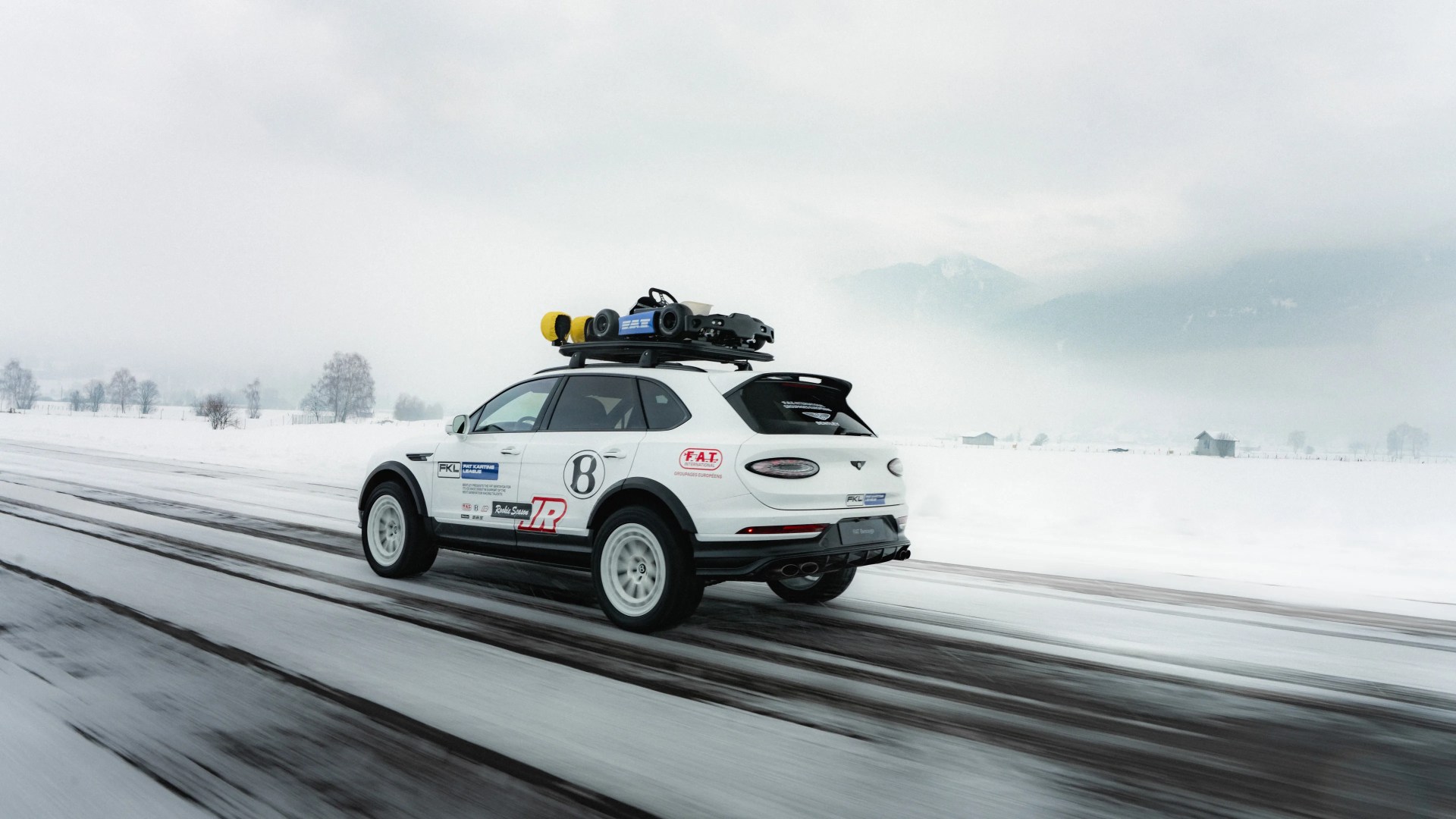 White Bentley SUV with racing decals and a go-kart on the roof driving on a snowy road.