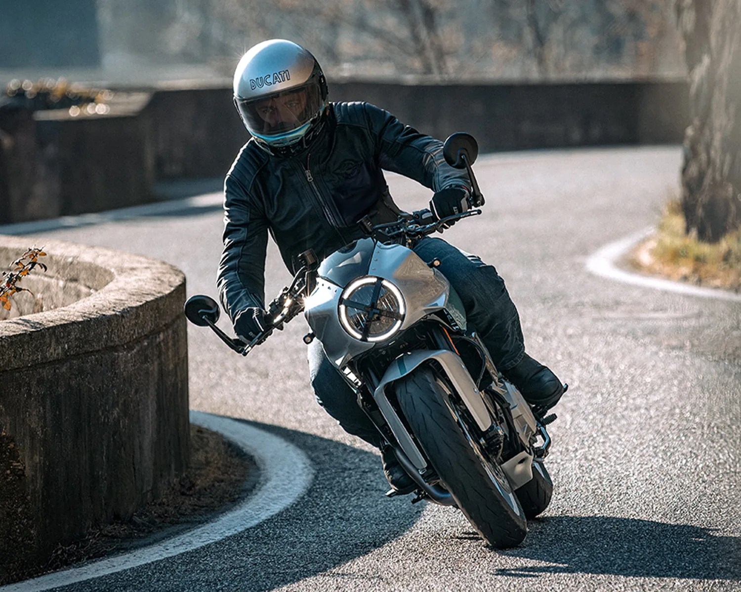 Motorcyclist wearing a silver Ducati helmet and black jacket leaning into a curve on a silver motorcycle.
