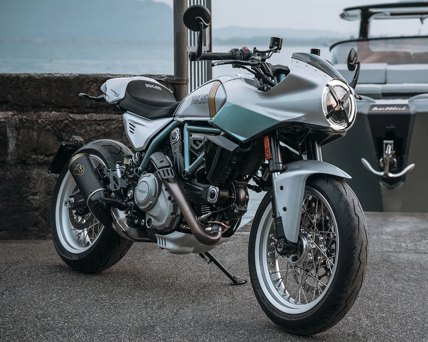 Silver and teal Ducati motorcycle with black seat and white rims parked near a stone wall and water.
