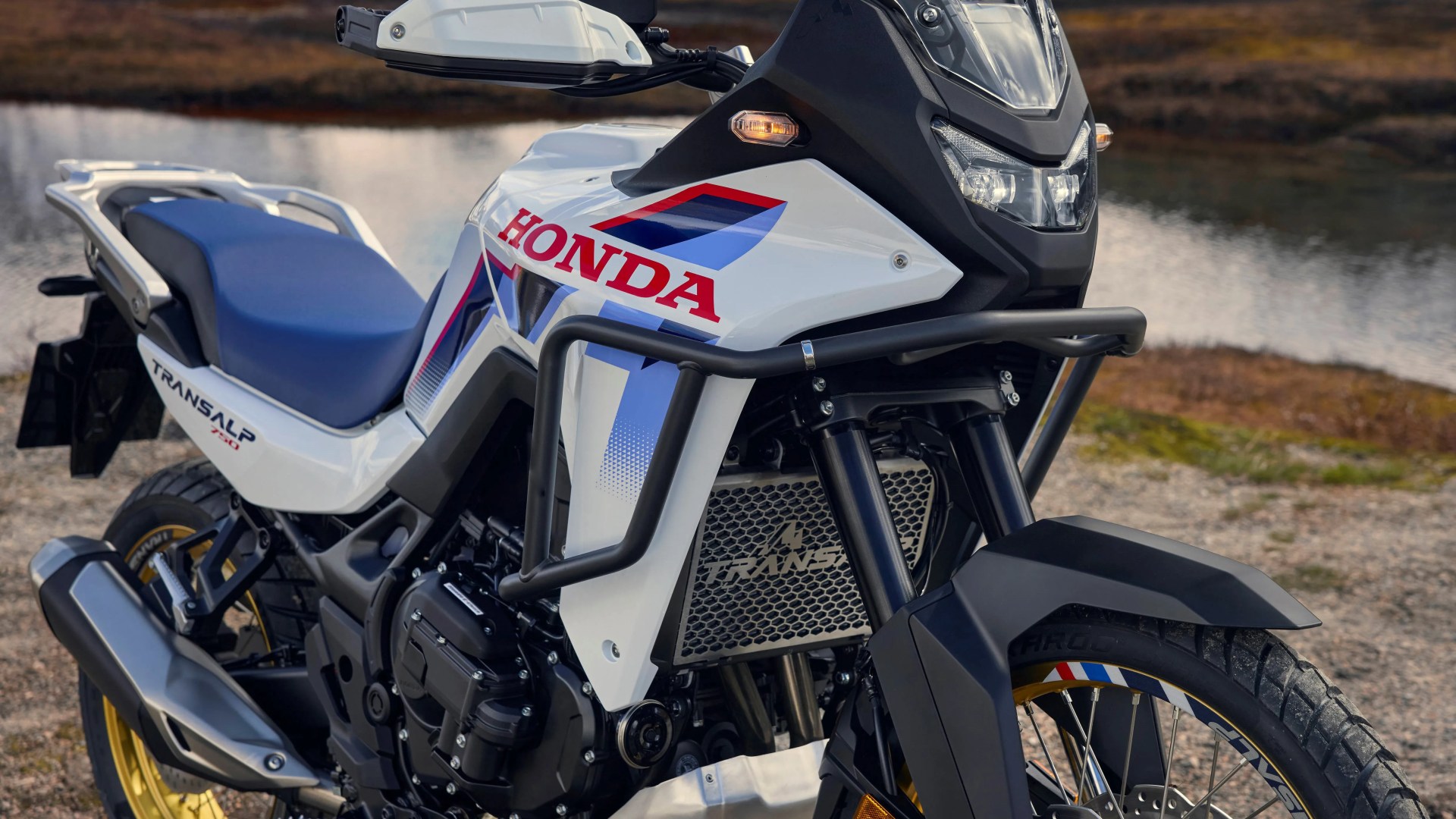 White Honda Transalp 750 motorcycle with blue seat and gold rims parked near a body of water.