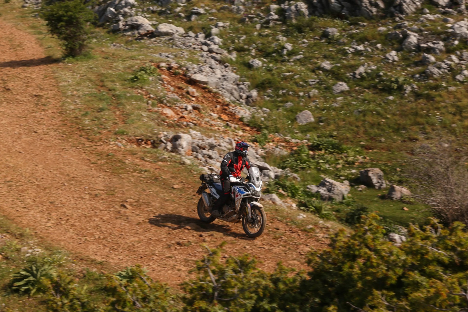 Motorcyclist in black and red gear riding a white and blue adventure bike on a rocky dirt trail.