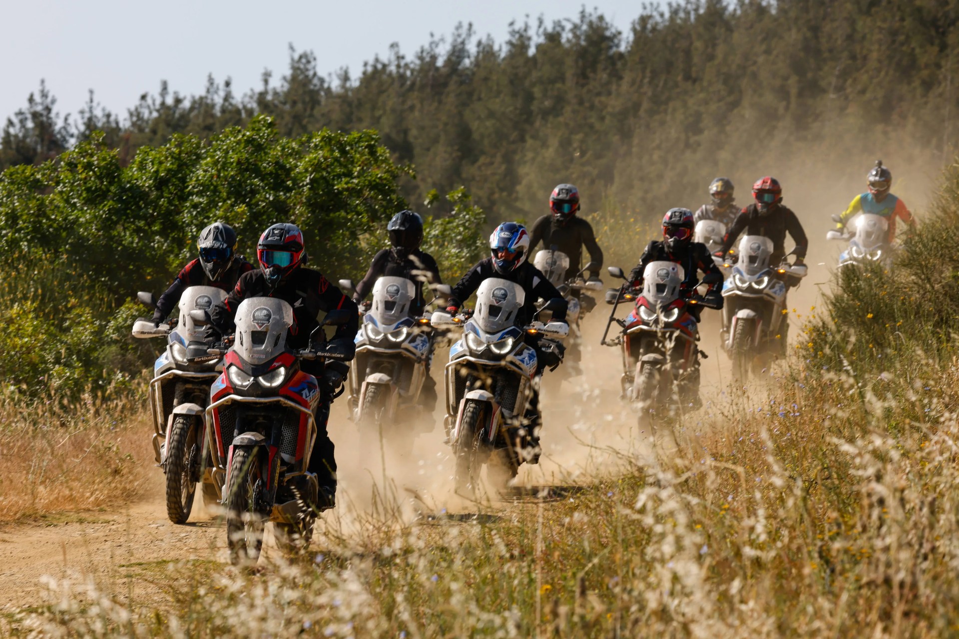 Group of motorcyclists riding adventure bikes on a dusty trail surrounded by greenery.