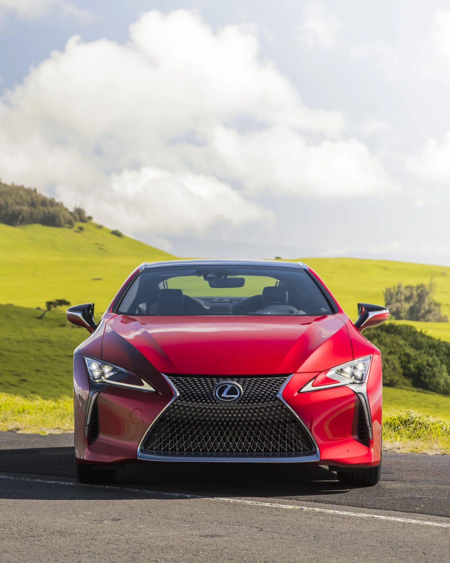Front view of a red Lexus sports car parked on a road with green hills and cloudy sky in the background.