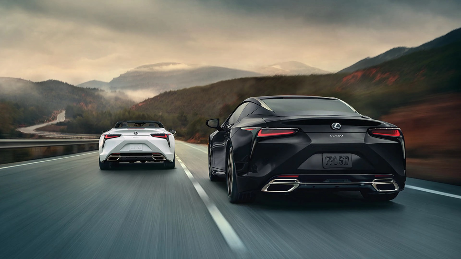 Two Lexus LC 500 cars, one black coupe and one white convertible, driving on a mountain road.