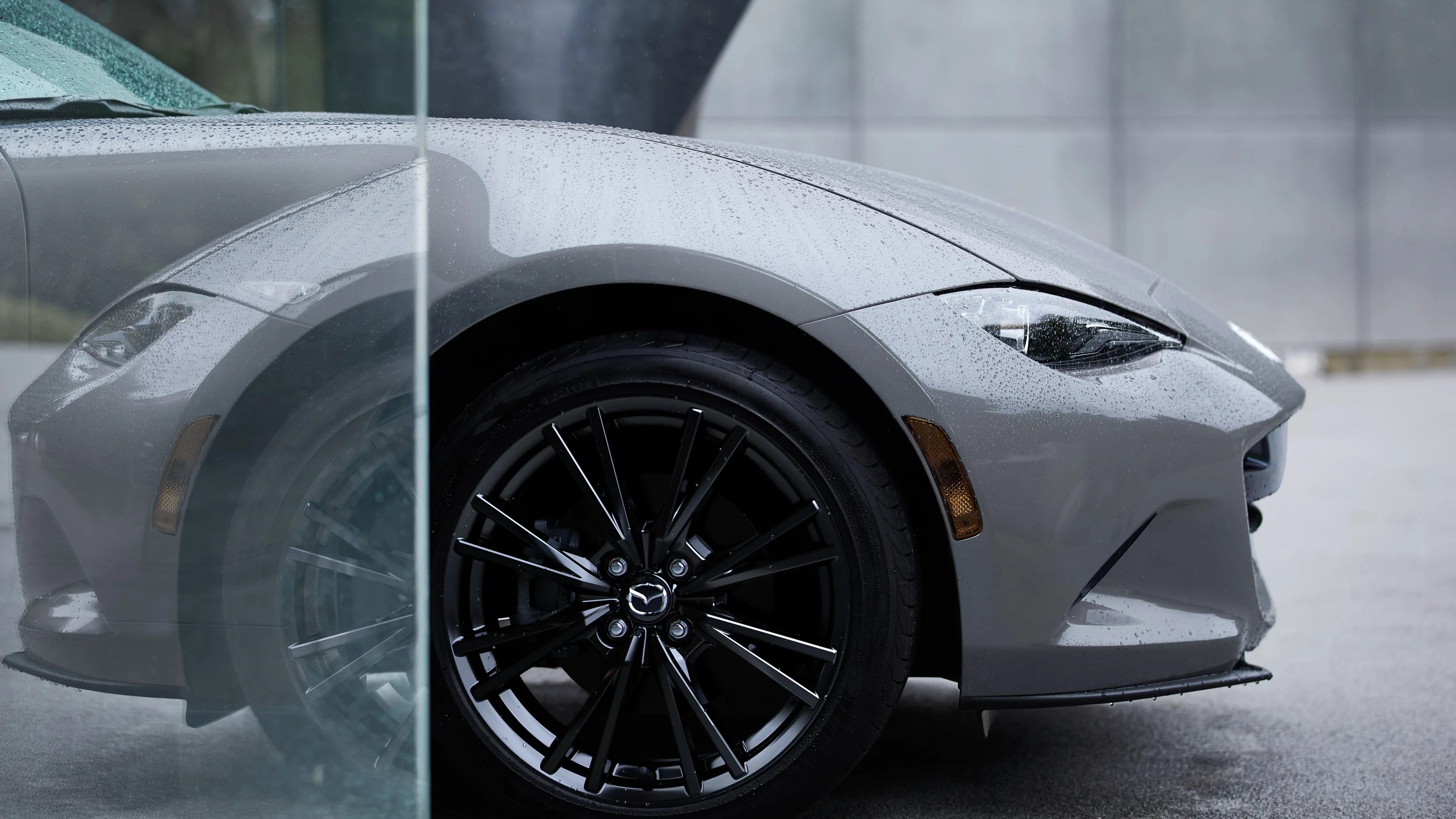 Front side of a gray Mazda car with rain droplets on the surface and black alloy wheel visible through a glass panel.