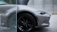 Front side of a gray Mazda car with rain droplets on the surface and black alloy wheel visible through a glass panel.