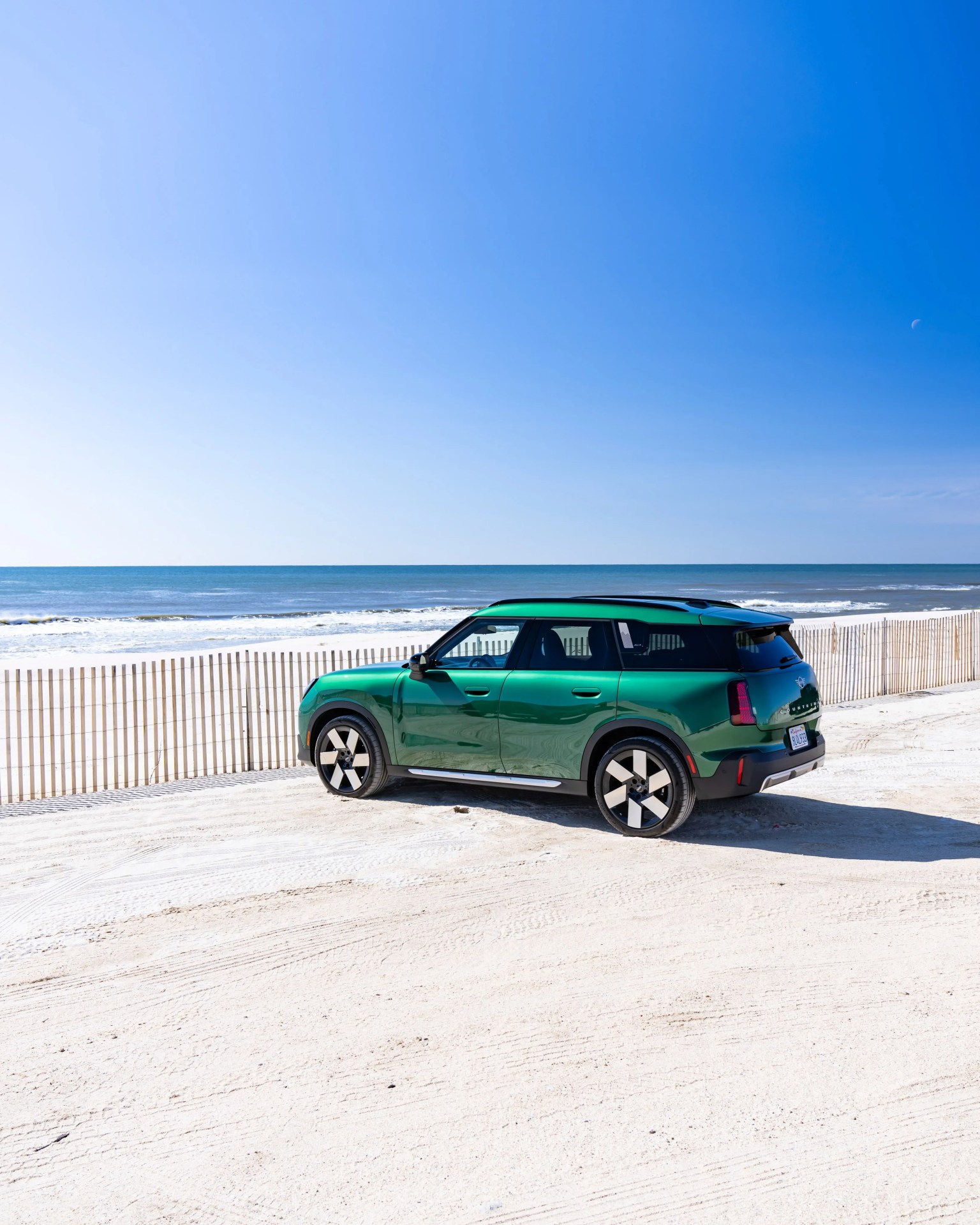 Green compact SUV parked on sandy beach near wooden fence with ocean and clear blue sky in background.