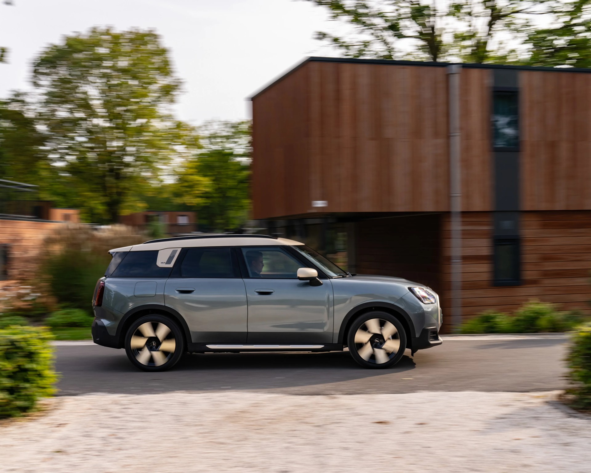 Gray compact SUV with black roof and beige side mirror caps driving past a modern wooden building.