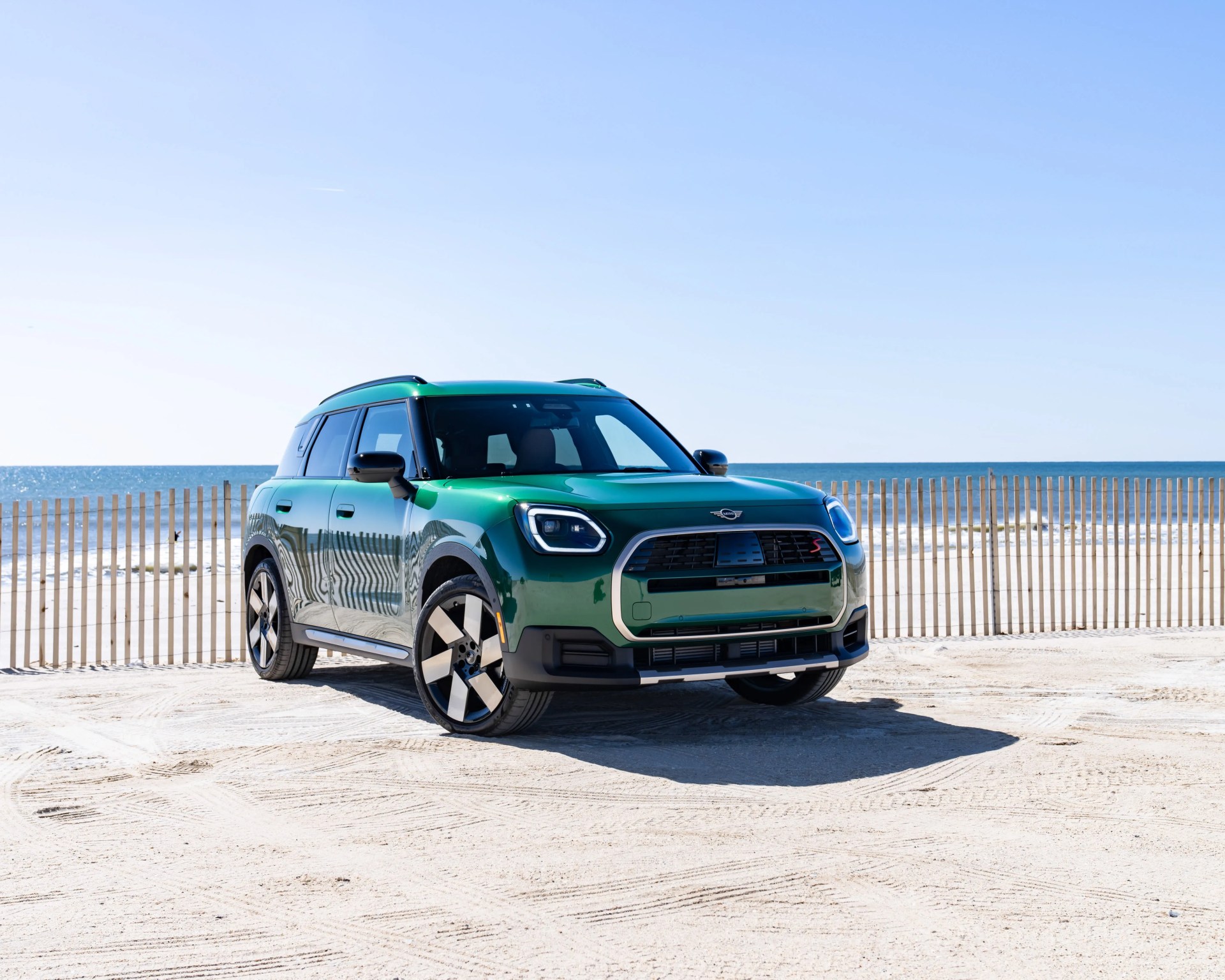 Green Mini Cooper SUV parked on sandy beach near wooden fence with ocean in background.
