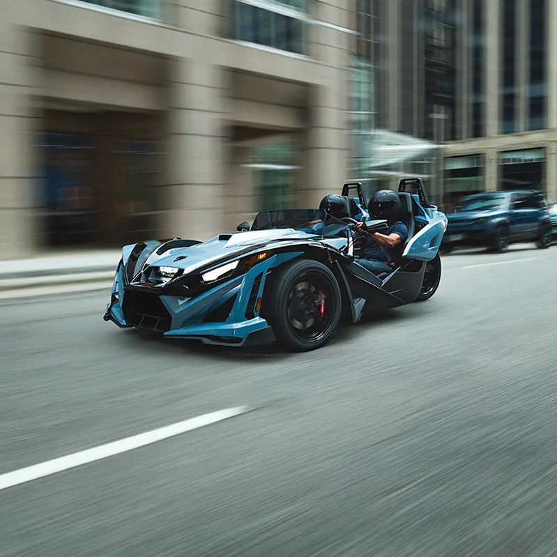 Blue and black three-wheeled open-top sports car with two passengers wearing helmets driving on a city street.