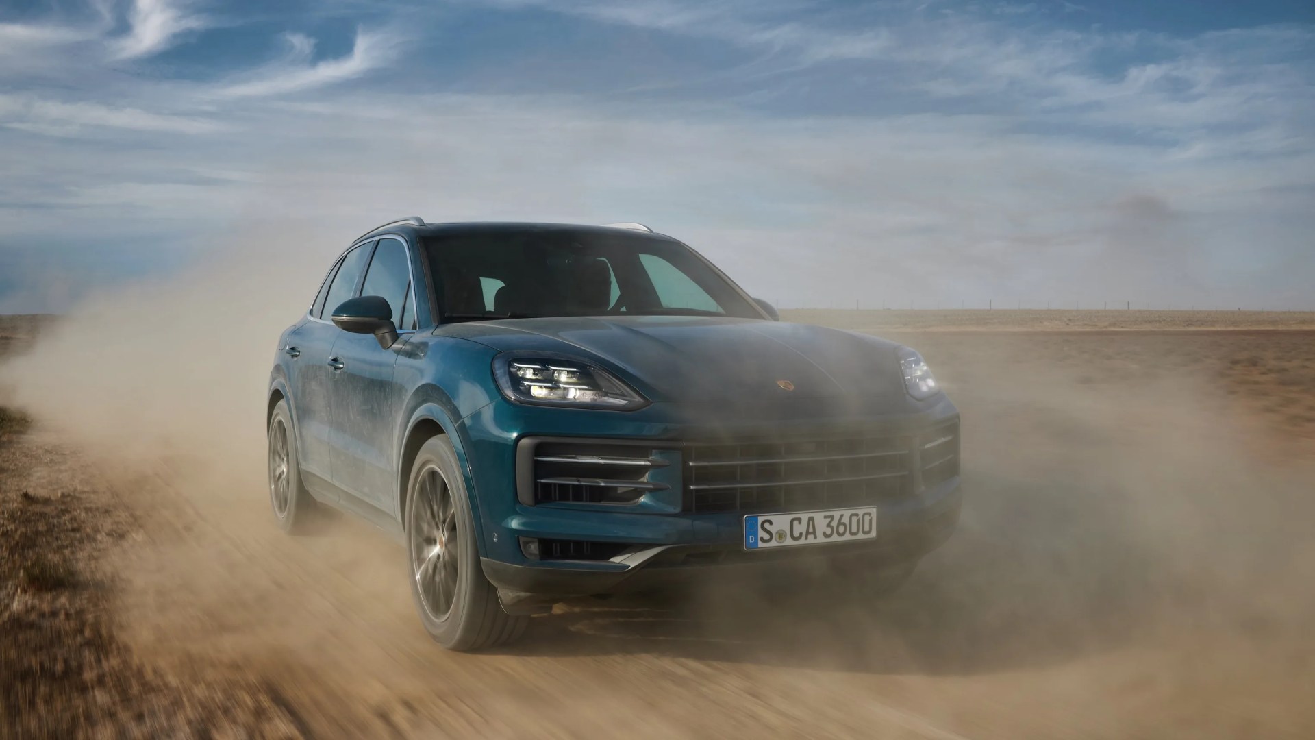 Blue Porsche SUV driving on a dusty dirt road with headlights on under a partly cloudy sky.