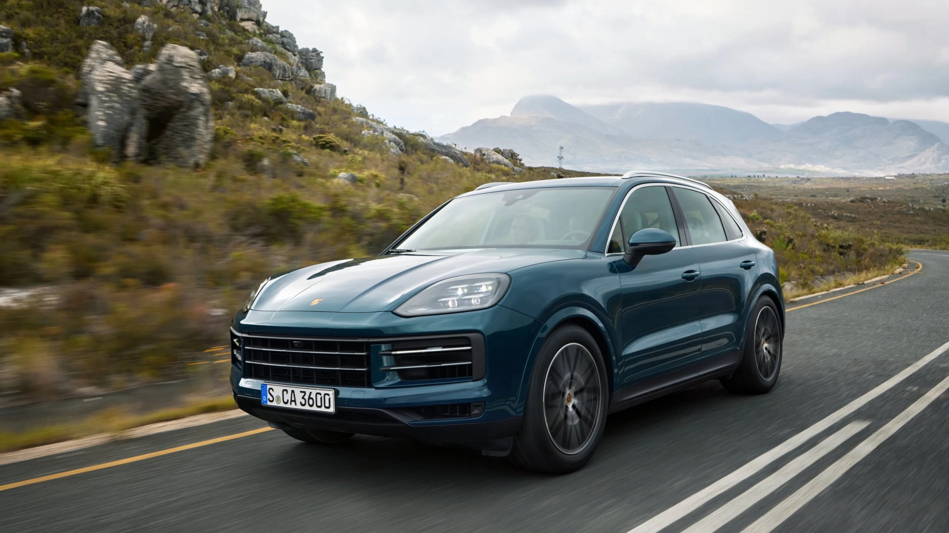 Blue Porsche SUV driving on a winding road with rocky hills and mountains in the background.